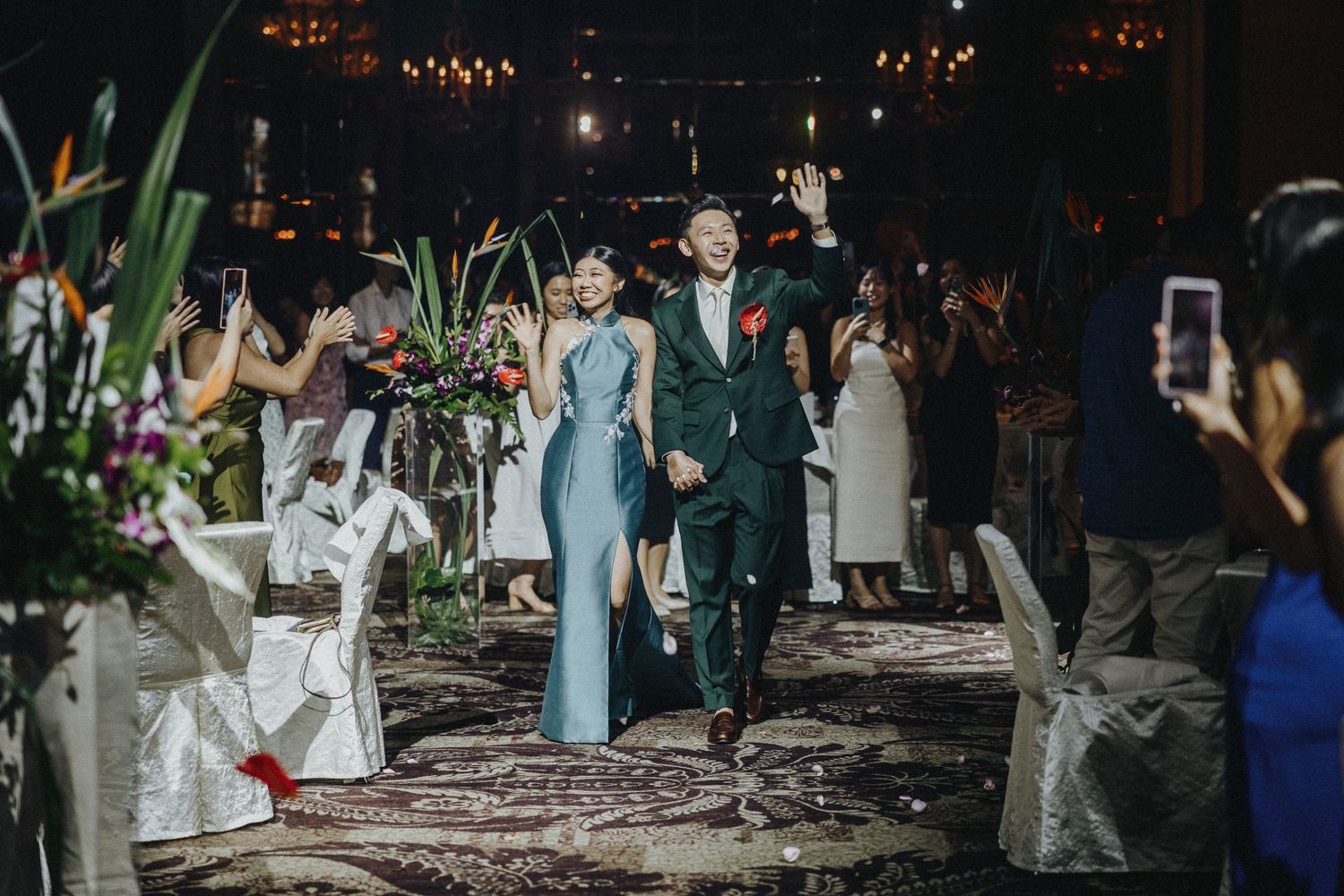 Wedding couple walking down aisle with floral decorations and guests capturing moment on phones.
