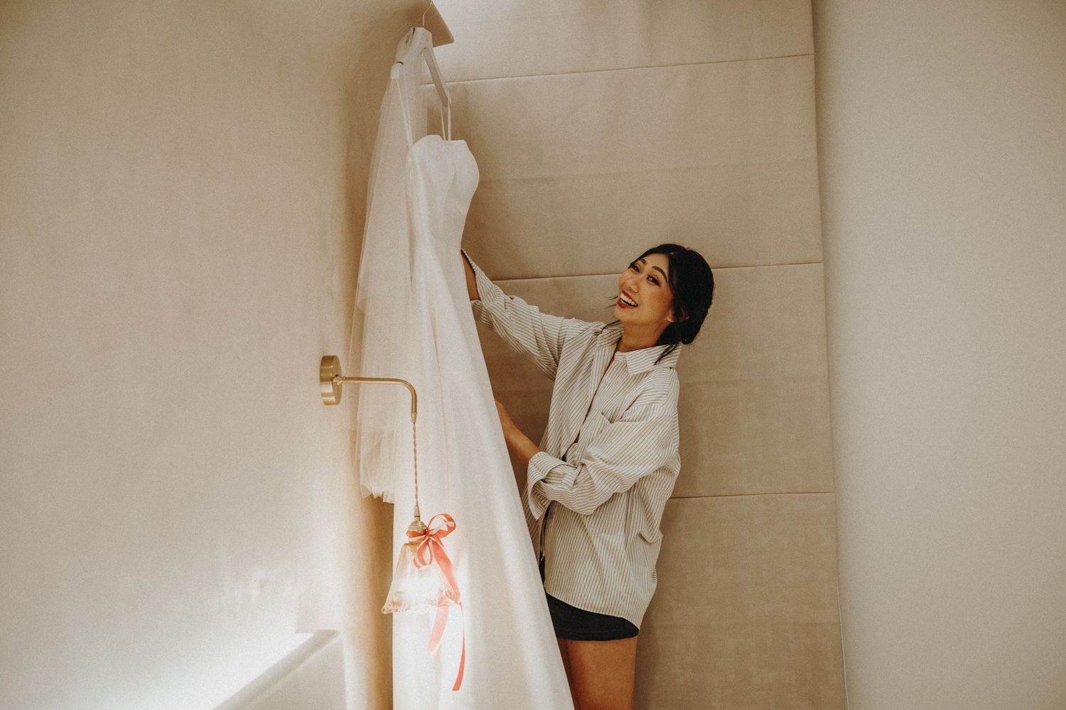 A wedding dress hangs on display as someone carefully inspects its pristine white fabric in a warmly lit room.