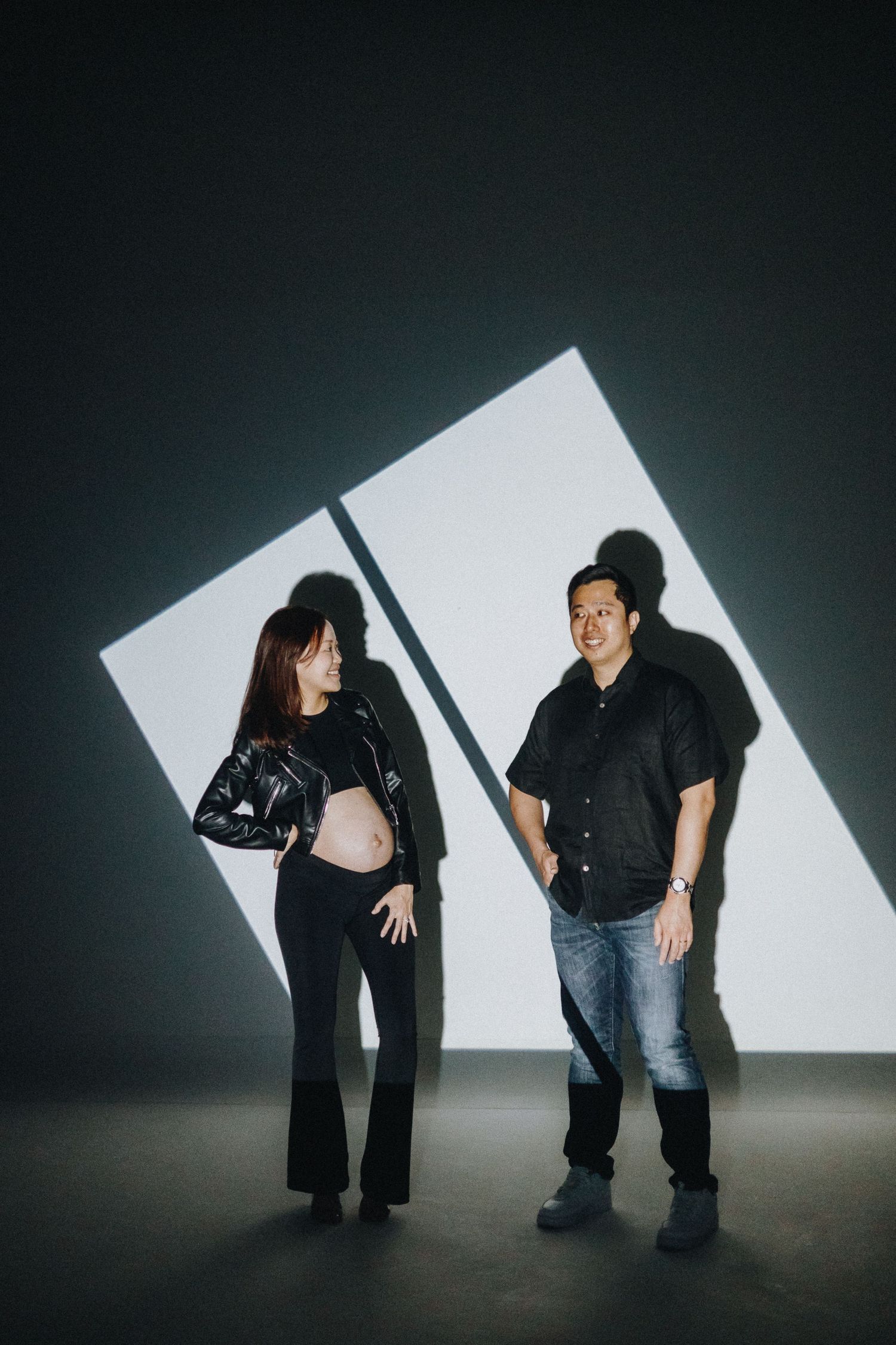 Maternity photoshoot with dramatic lighting featuring couple in black outfits against geometric shadows.