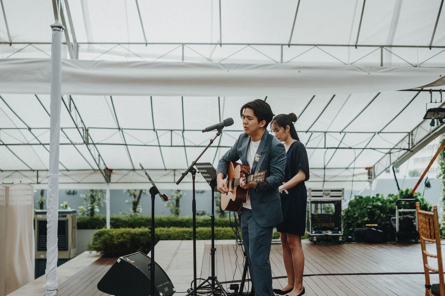 Musicians perform on stage at an outdoor rooftop venue with white canopy and gardens in background.