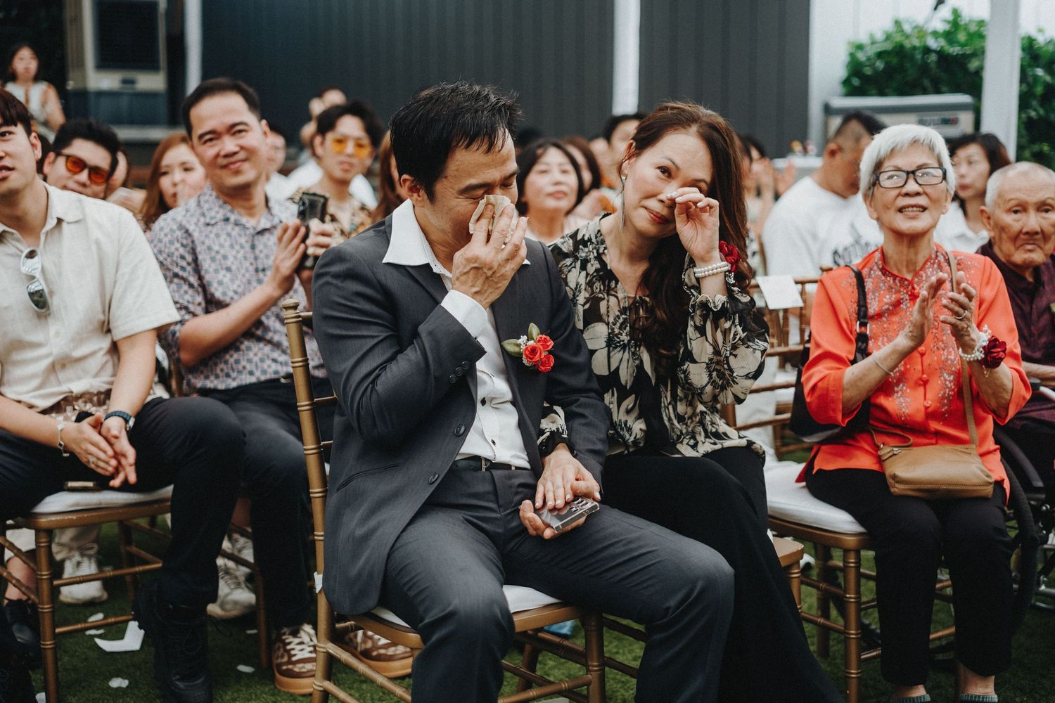 Guests at an outdoor wedding ceremony show emotional reactions while sitting in wooden chairs.