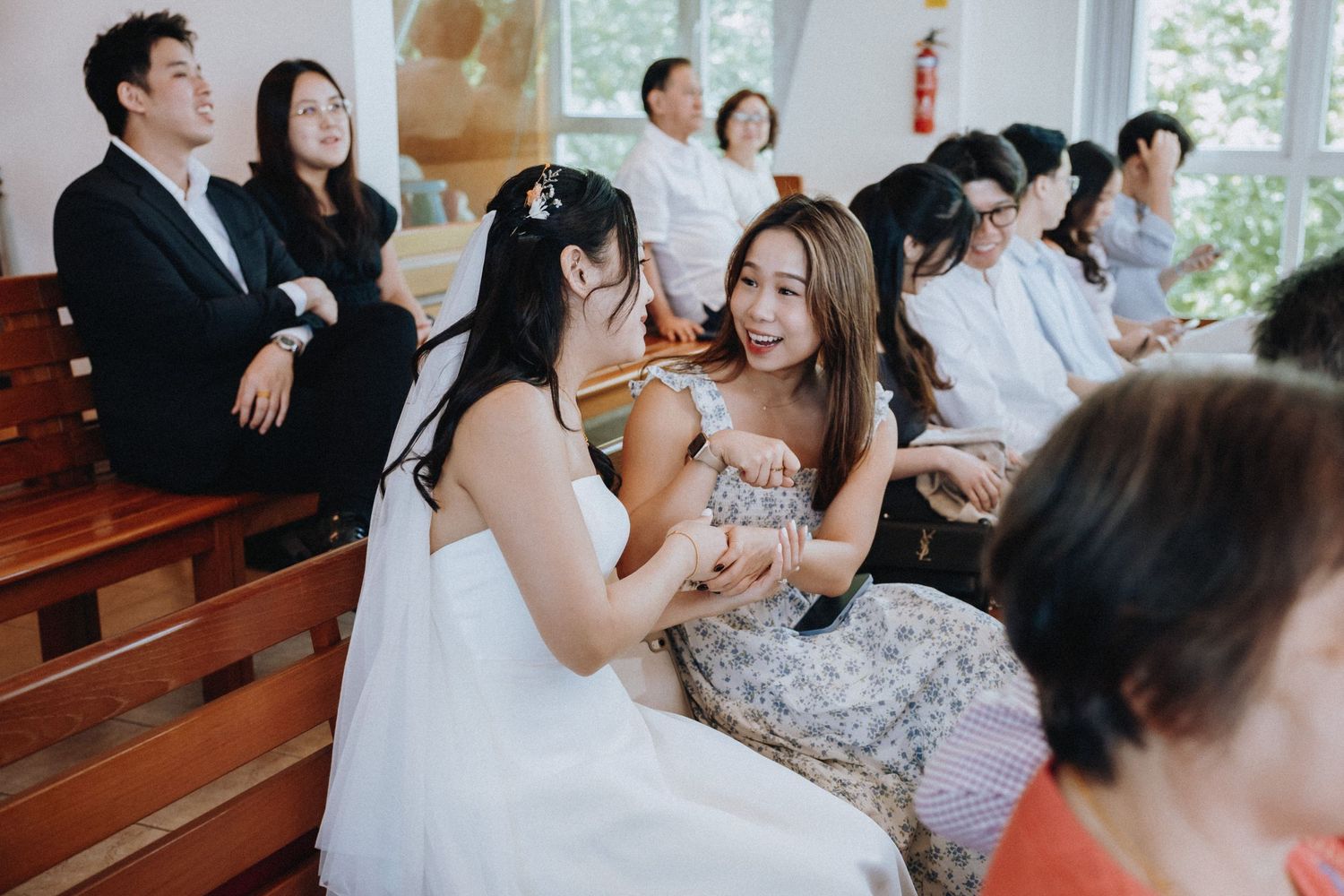 Bride laughing with female guest seated in church pews during joyful wedding ceremony celebration.