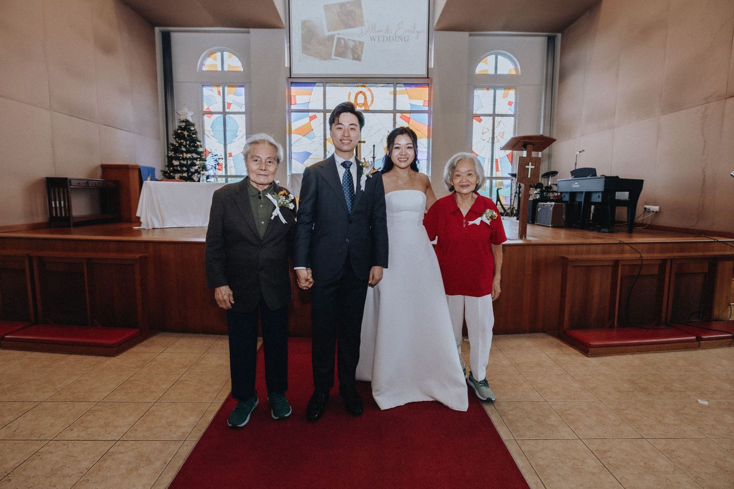 Bride and groom pose with two elderly family members on a red carpet inside a church after the ceremony.