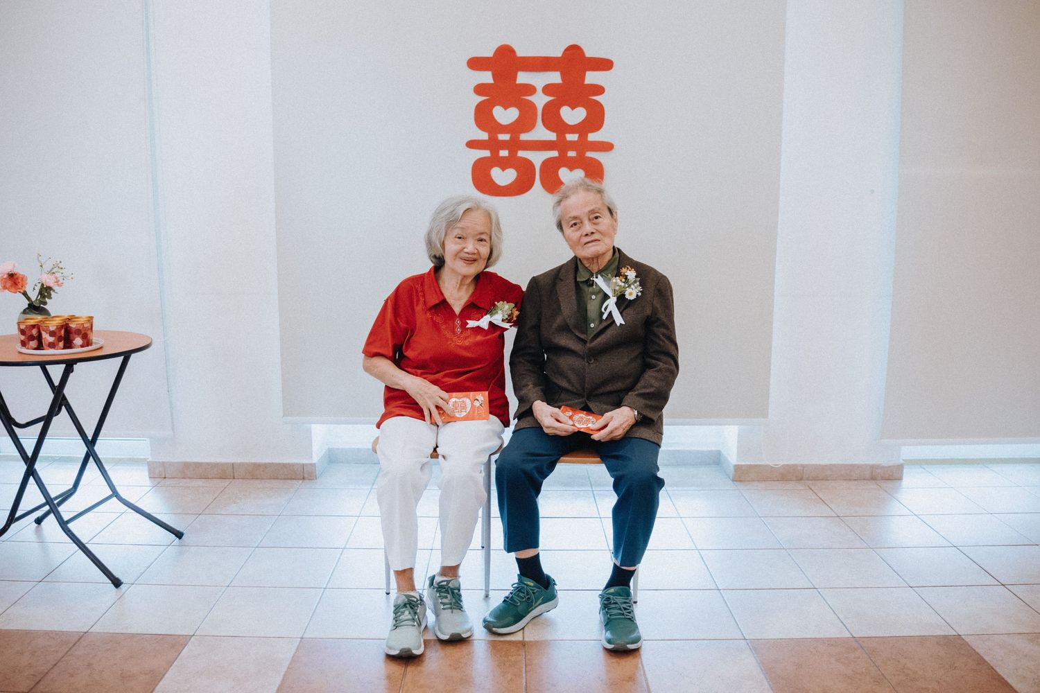 Elderly couple sits together in front of a red double happiness symbol at a wedding.