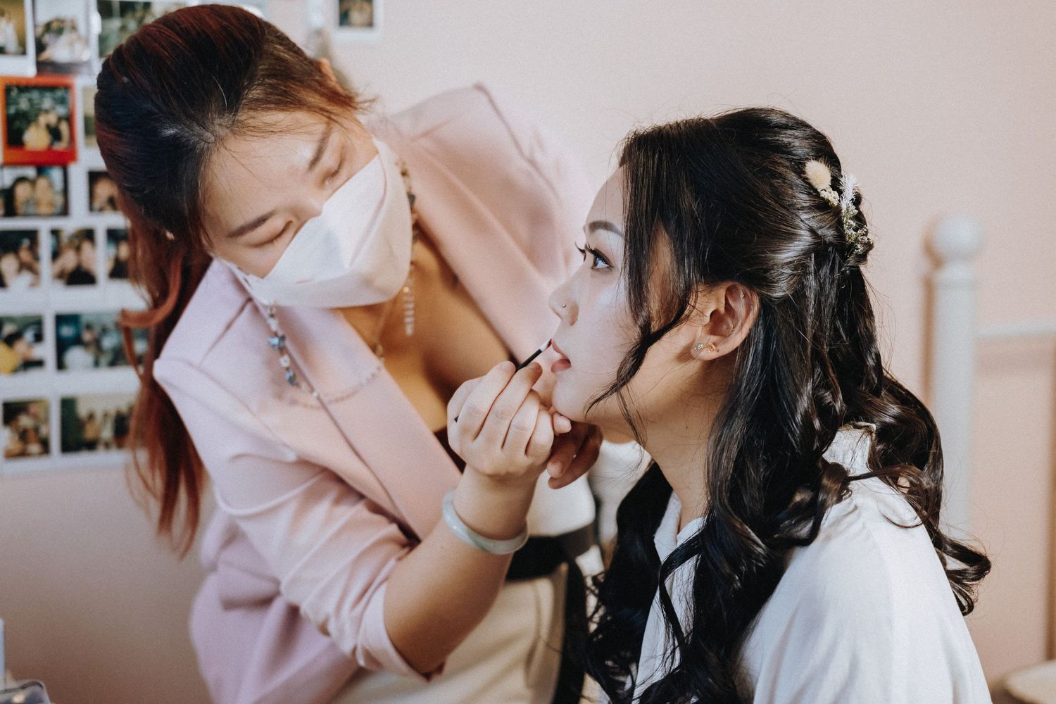 A makeup artist wearing a face mask carefully applies lipstick to a young woman with elegant hair accessories.