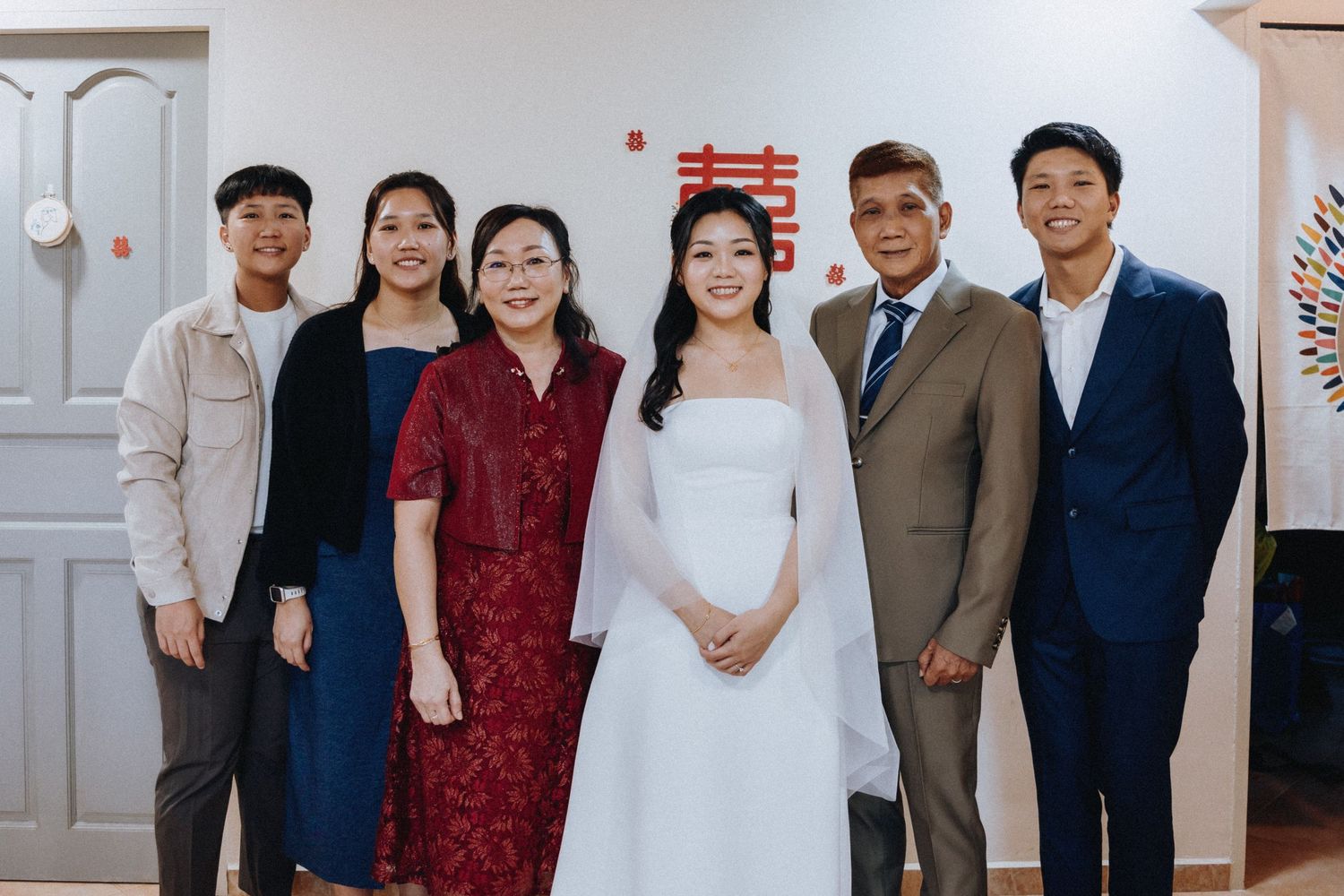 A bride in a white dress poses with her family at a wedding celebration, with a red double happiness symbol on the wall.