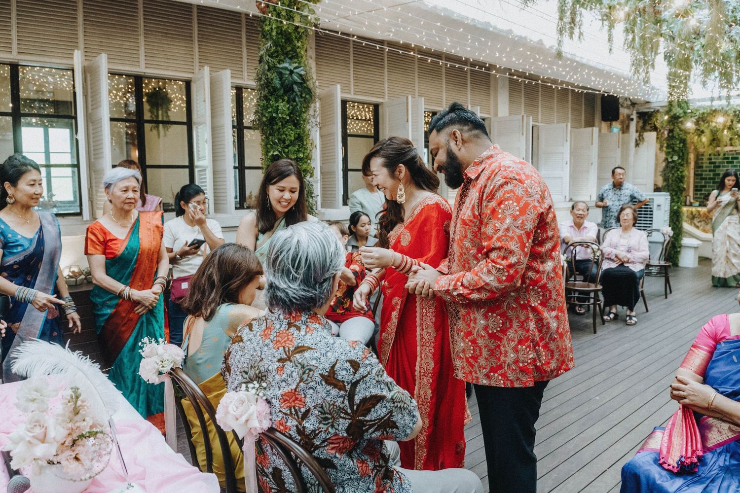 Guests in colorful traditional attire gather outdoors during a multicultural wedding ceremony on a decorated wooden deck.