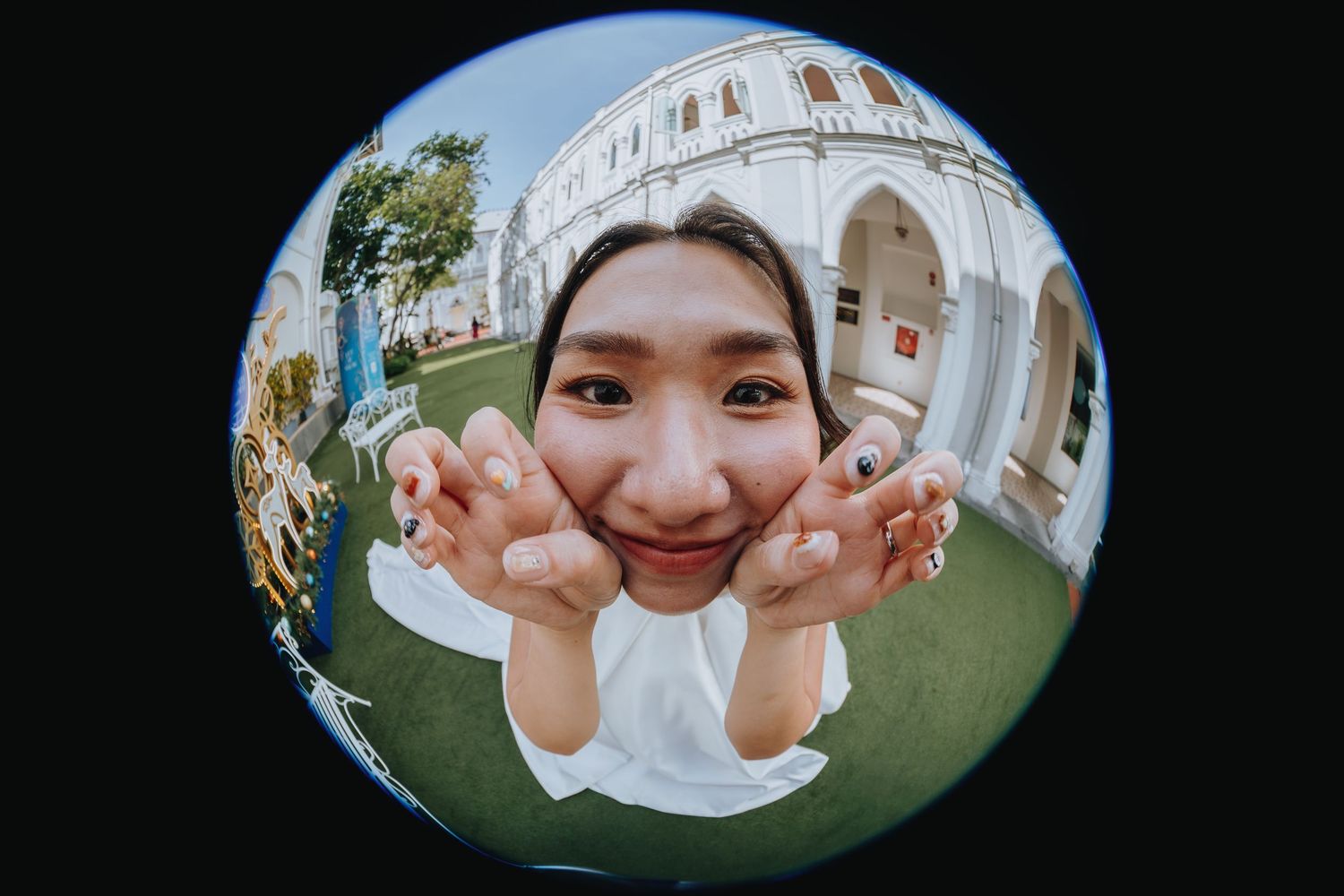 Fun fisheye lens portrait of a smiling young woman posing outdoors near a white colonial building.