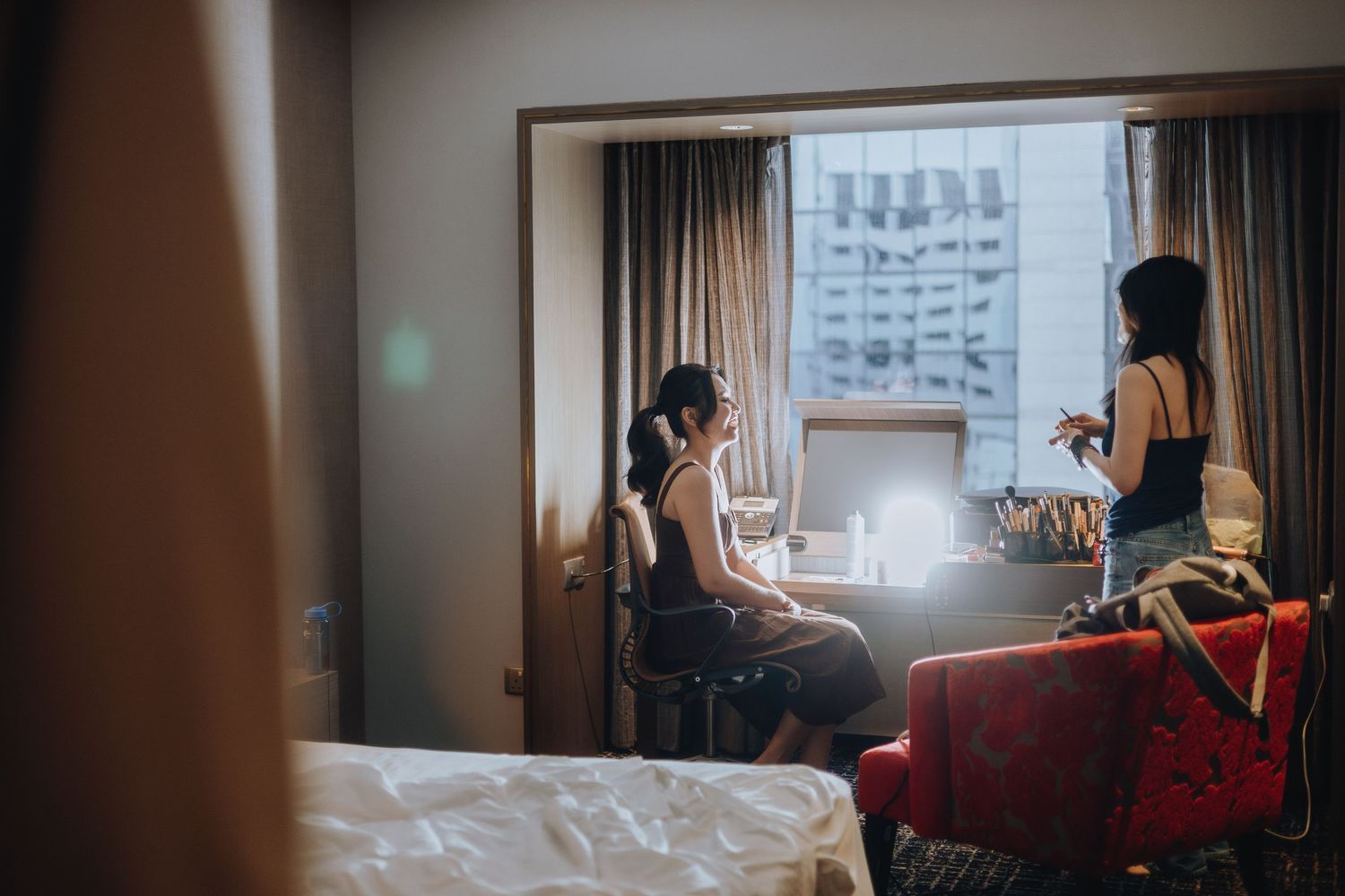 Two women in a hotel room, one seated at a vanity mirror getting ready, the other standing nearby with luggage.