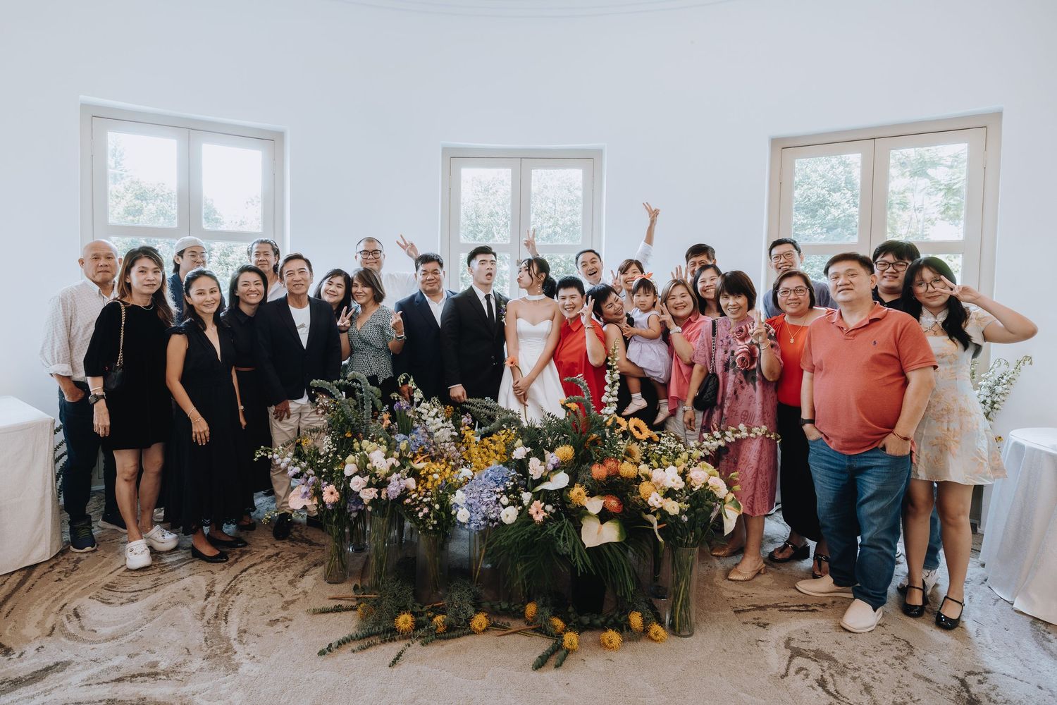 Wedding group photo with bride, groom, and guests gathered around large floral arrangement.