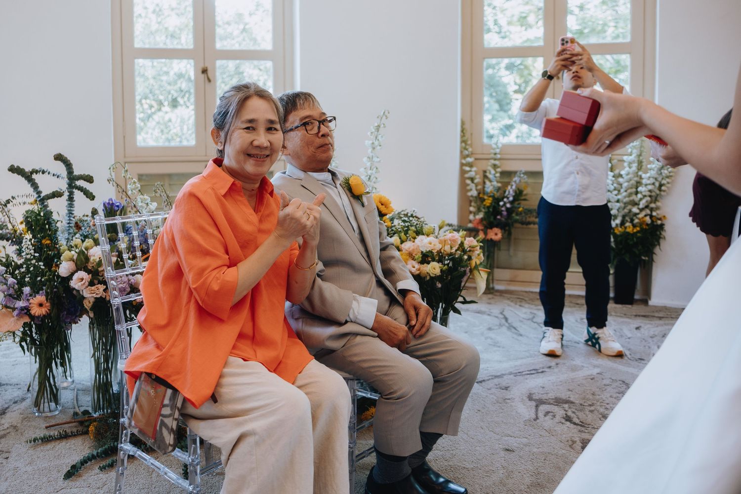 Elderly couple in orange and gray outfits smile during a wedding ceremony with flowers in background.