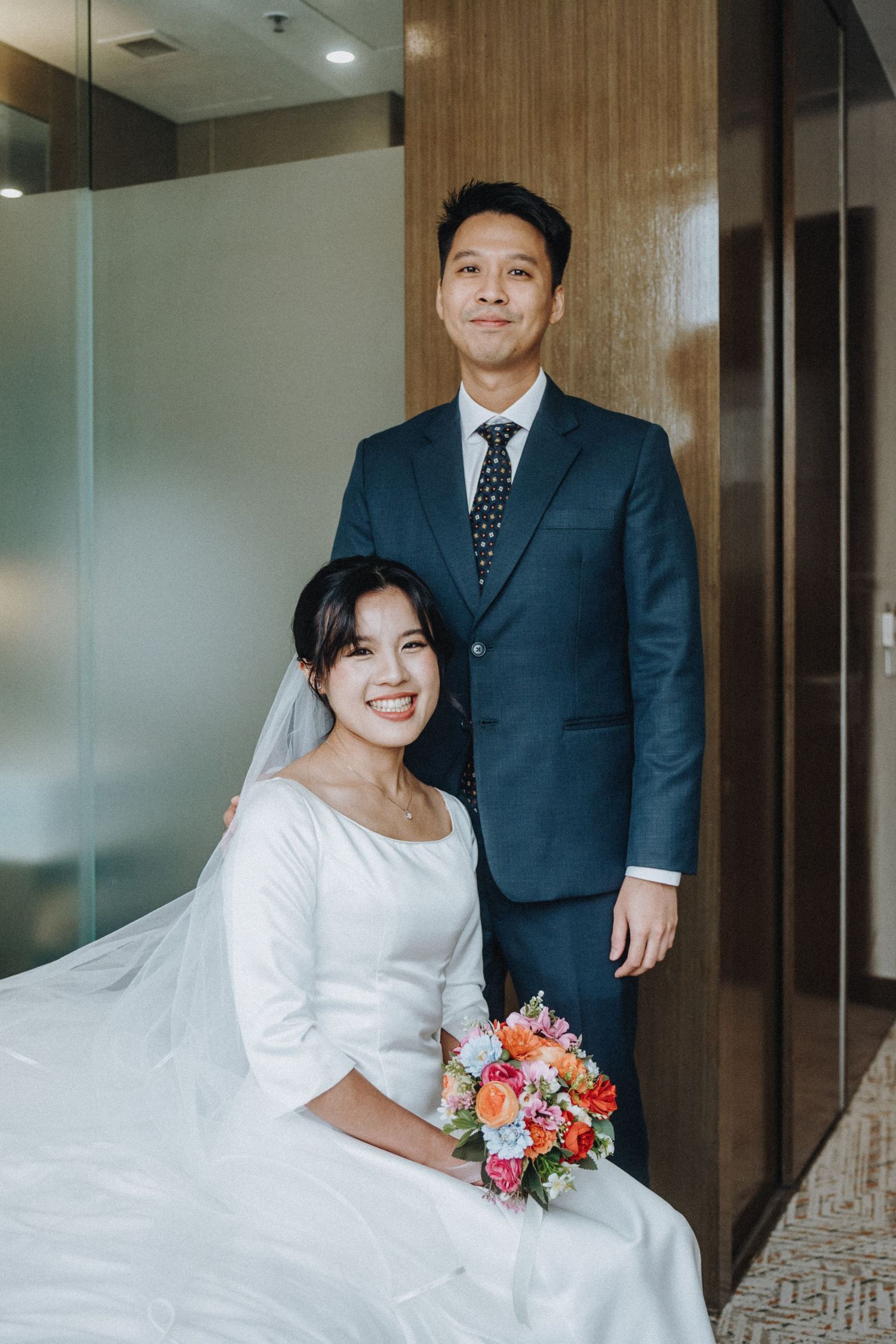 A smiling bride in a white wedding gown holding a colorful bouquet sits beside a groom in a navy suit.