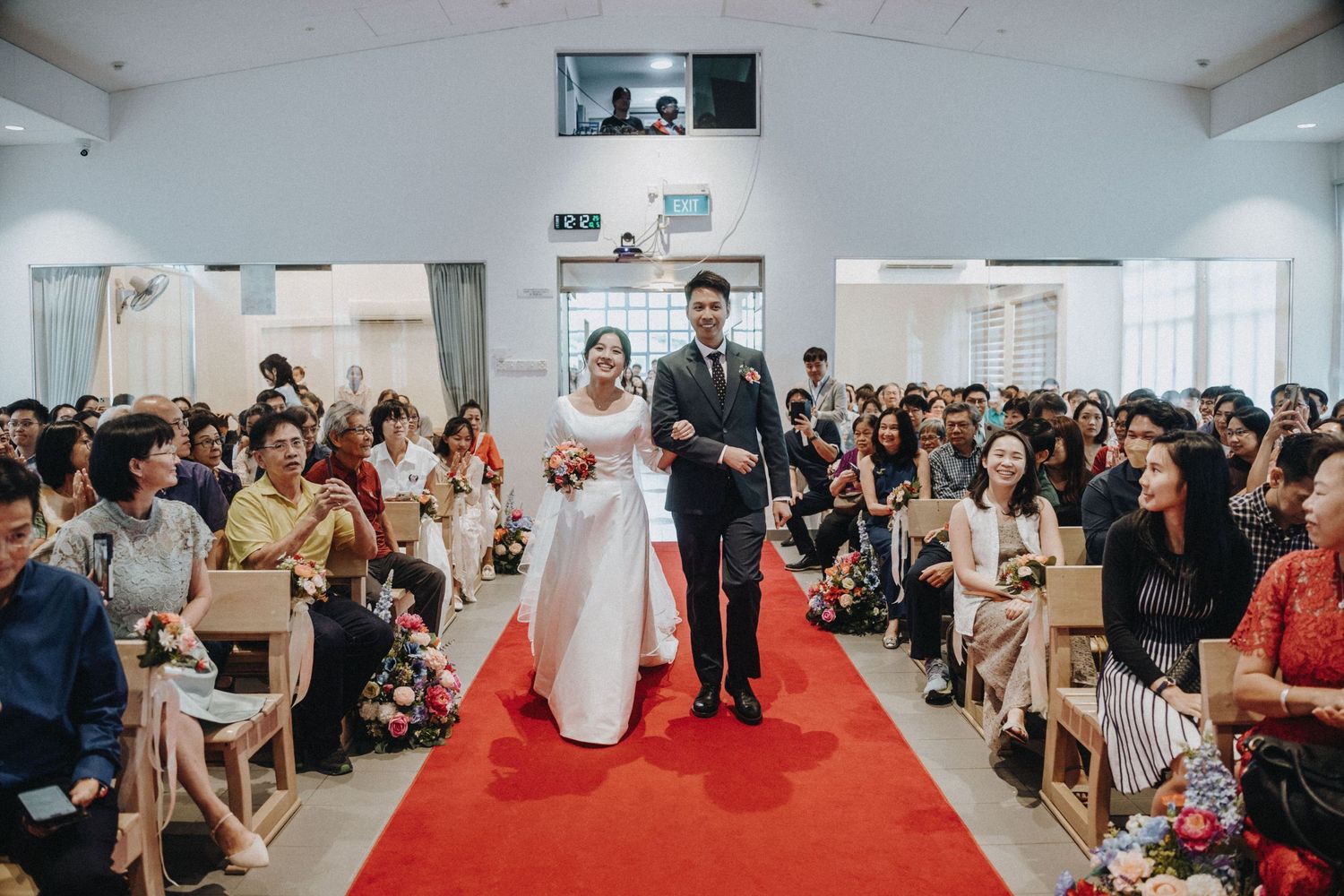 A bride and groom walk down a red-carpeted church aisle surrounded by seated wedding guests.