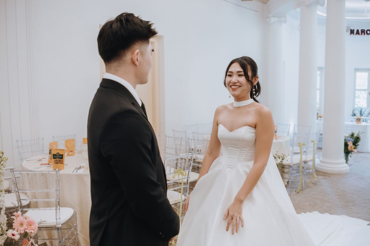 A smiling bride in a white ball gown faces her groom in a black tuxedo during their elegant indoor wedding ceremony.