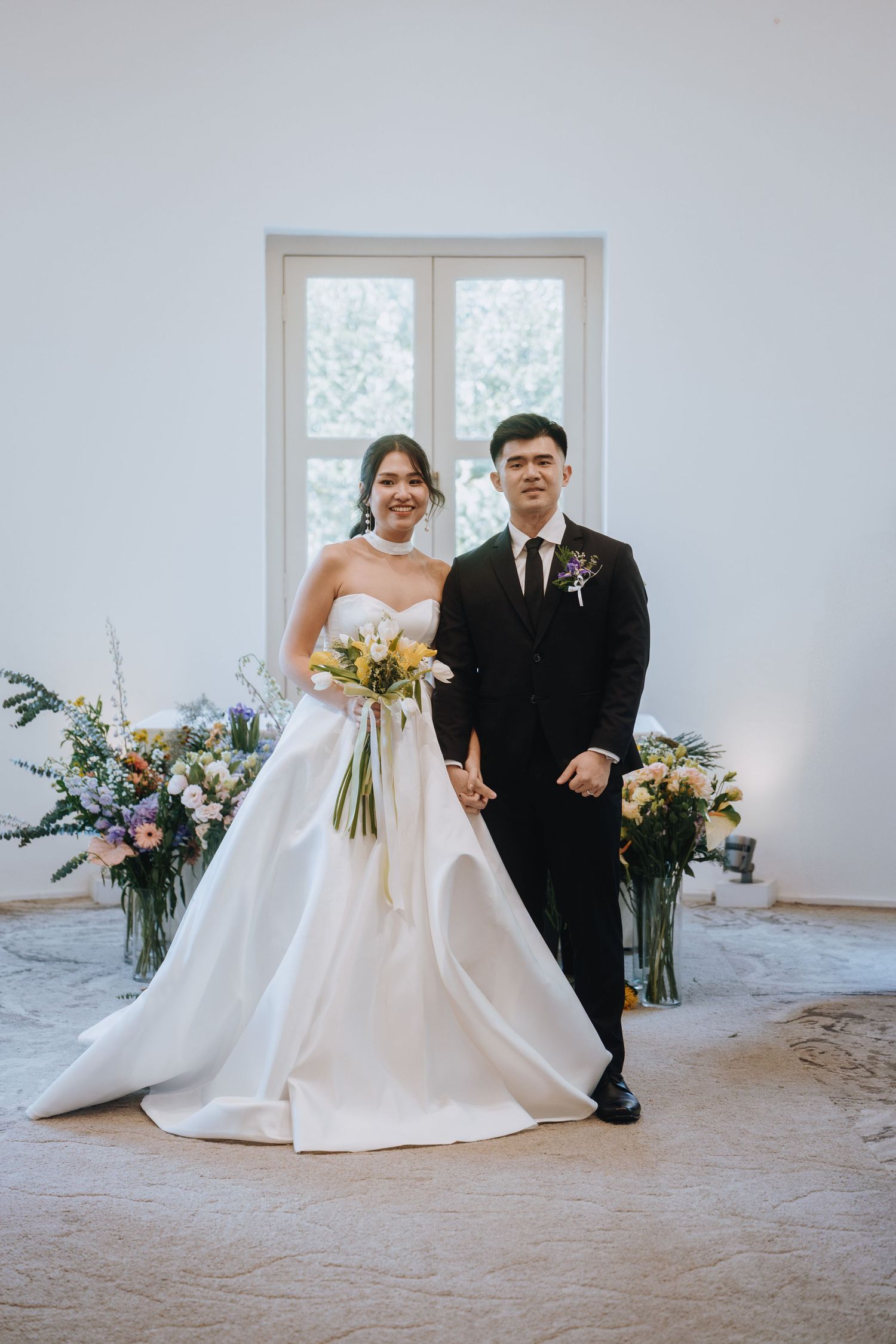 Bride holding yellow and white bouquet stands with groom in black suit before large window with florals.