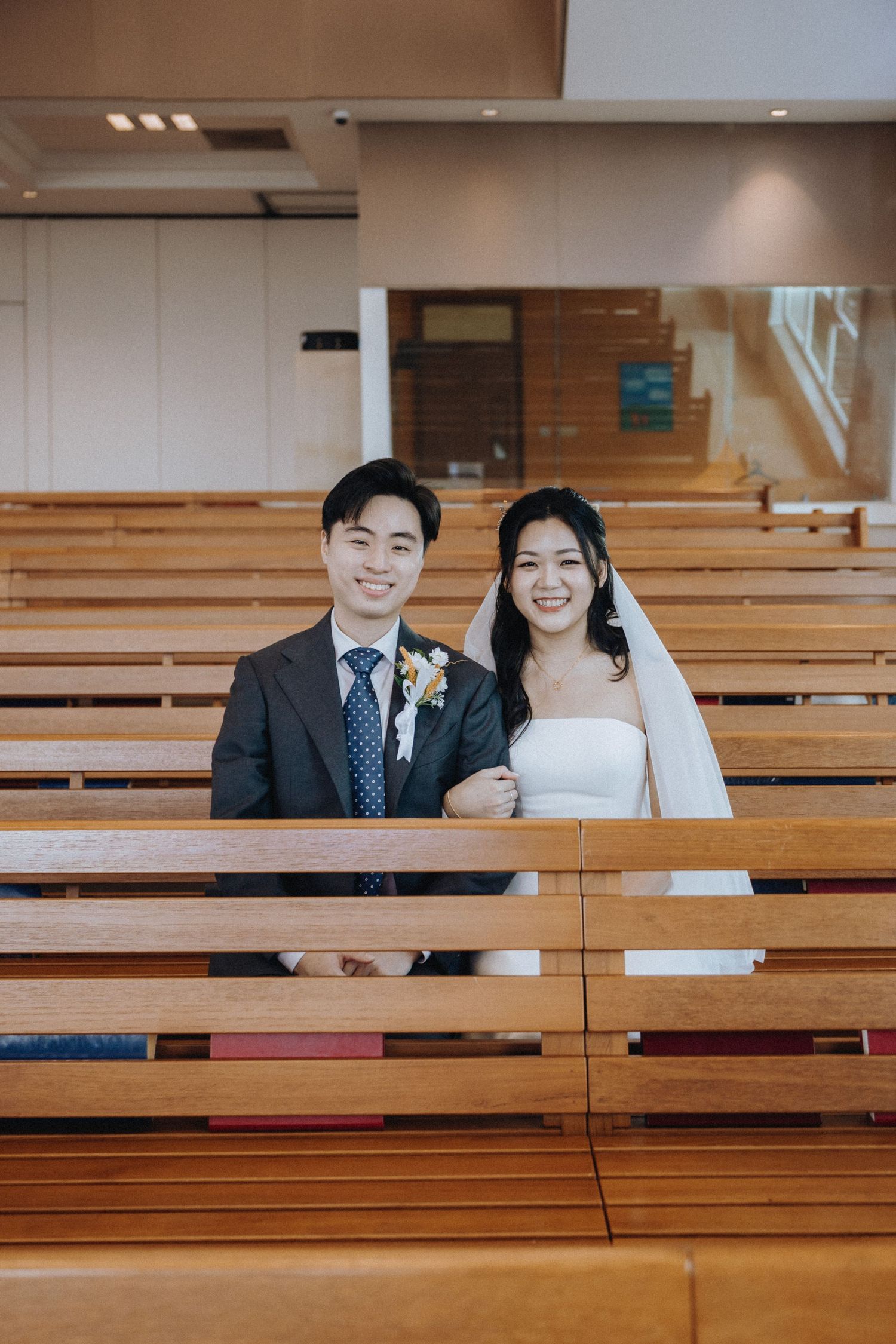 Happy couple in wedding attire sitting together among wooden church pews.