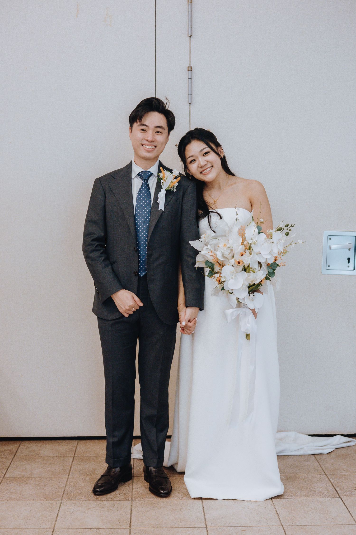 A smiling bride and groom pose together, she holds a white floral bouquet in a strapless gown, he wears a dark suit with blue tie.