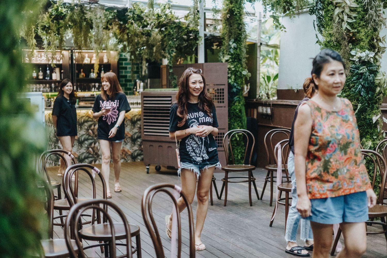 Women walking through a lush garden cafe with wooden chairs and green plants in a tropical setting.
