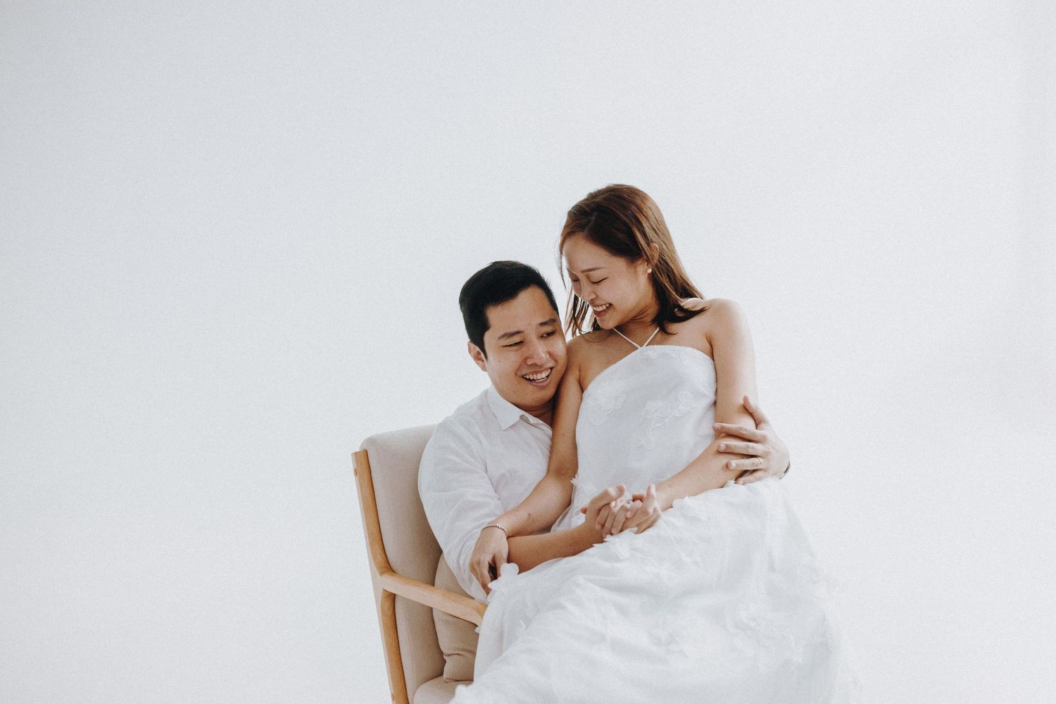 Two people share a joyful moment while sitting together in white attire against a minimal white backdrop.