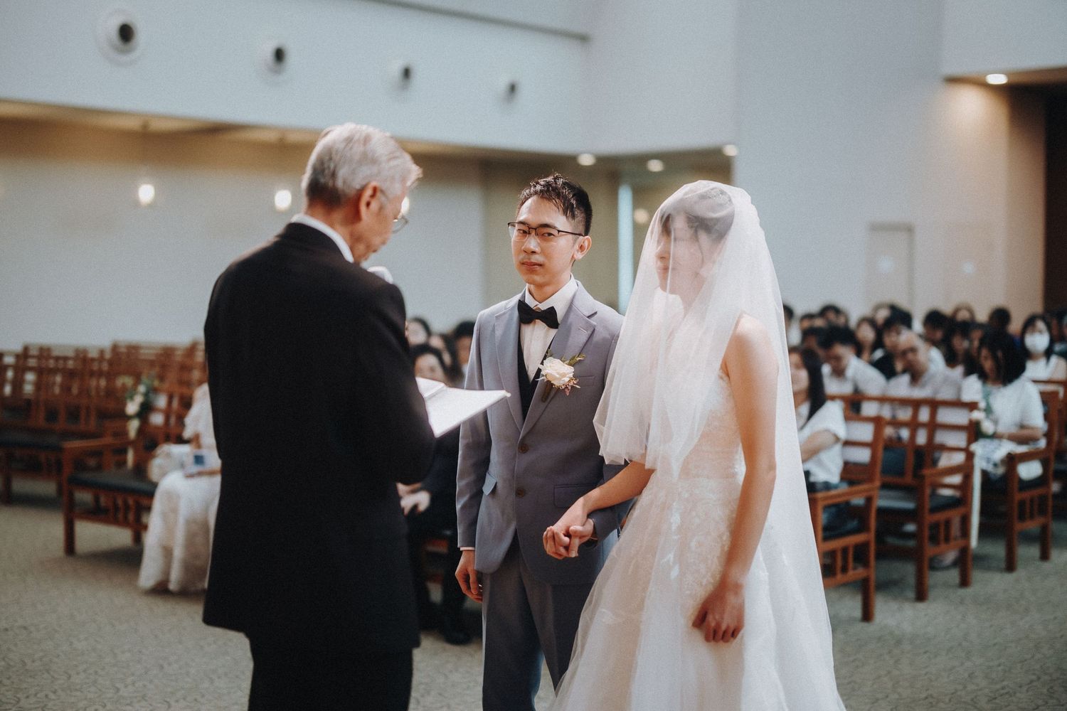 Wedding ceremony taking place in a bright, minimalist church hall with wooden chairs and white walls.