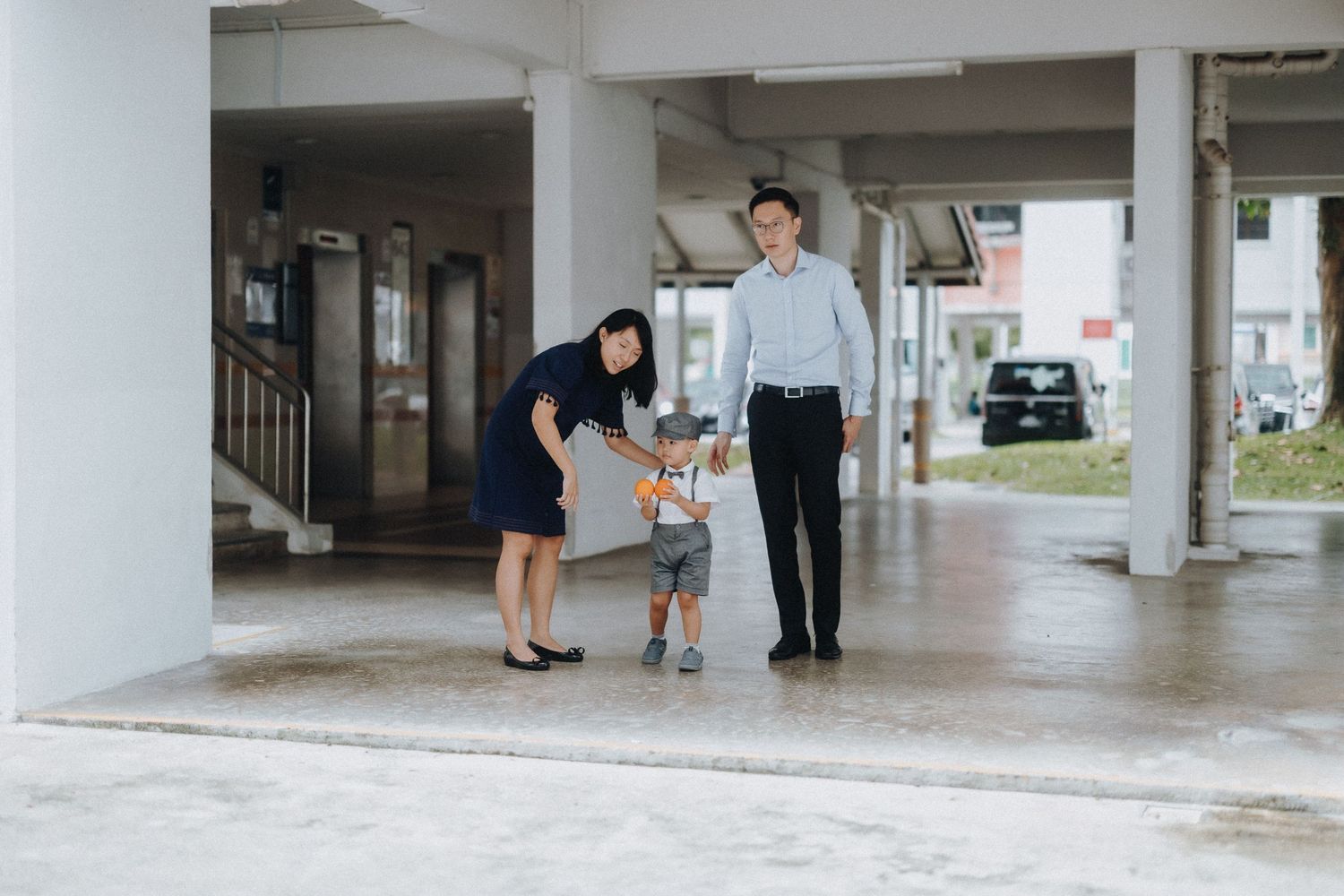 A family walks together beneath concrete pillars in an open covered area with parked cars visible in background.