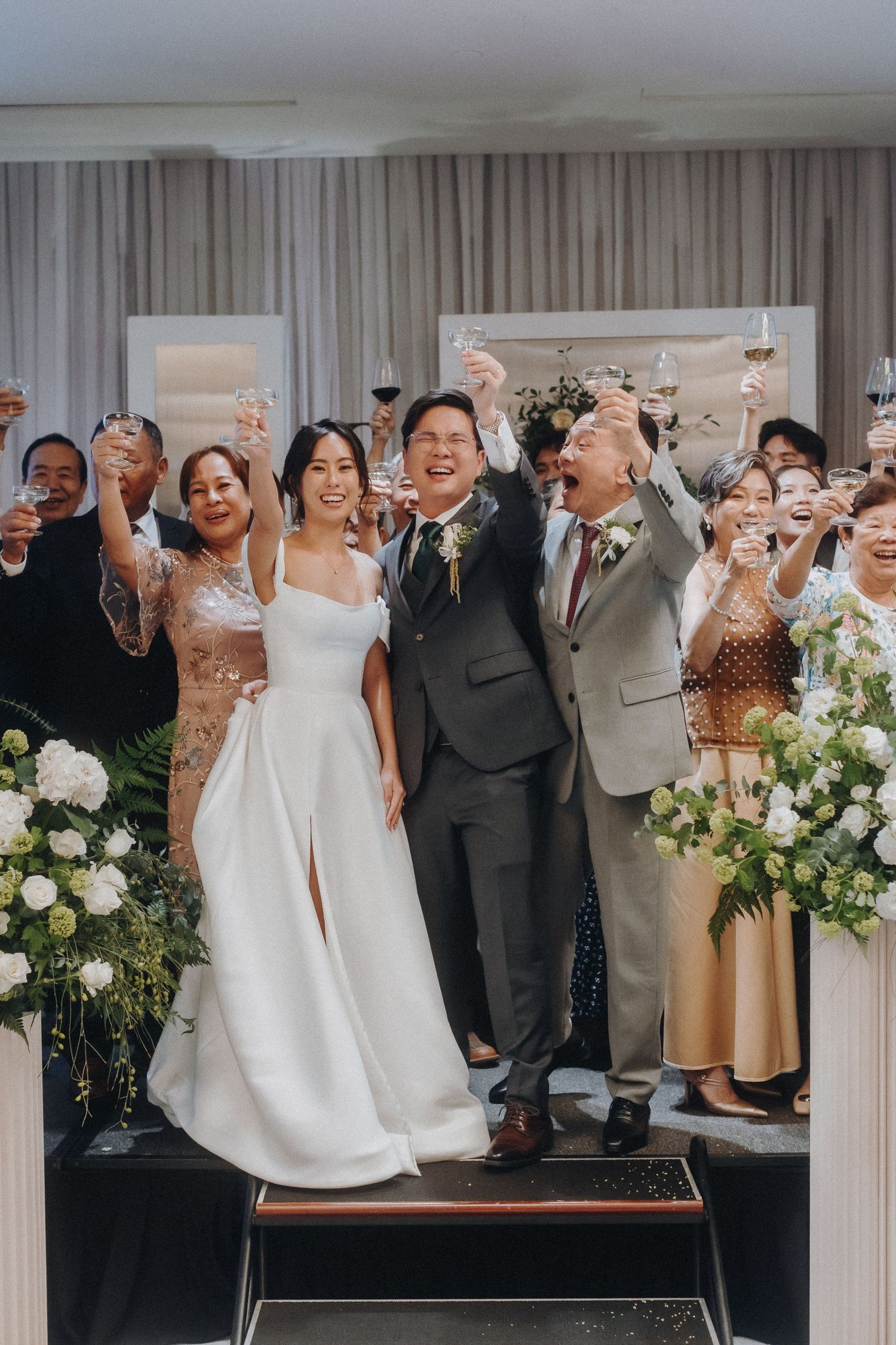 Wedding celebration with guests toasting against a backdrop of white flowers and drapes.