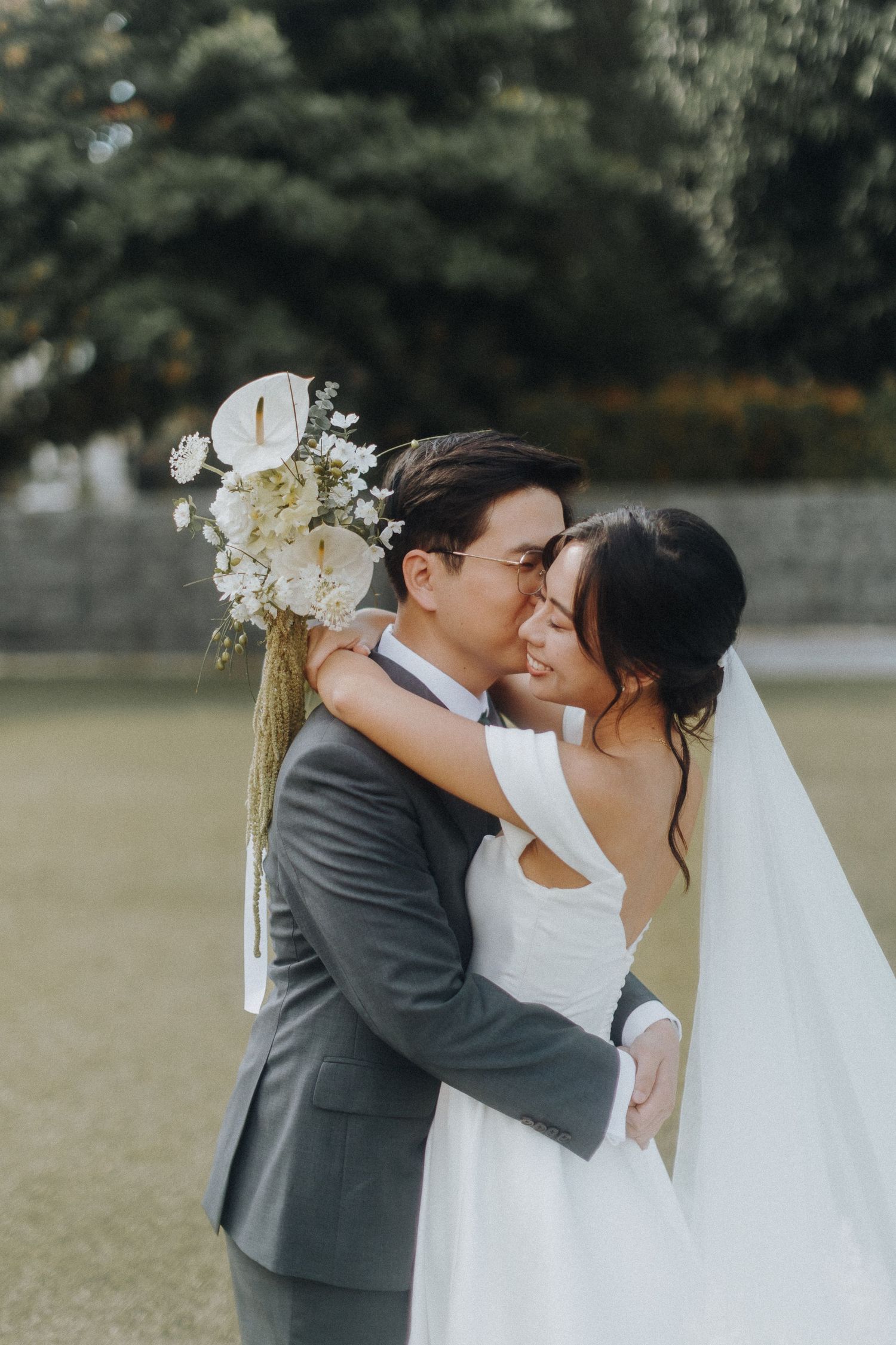 Newlyweds share a romantic kiss during their outdoor wedding photoshoot with white floral decorations.