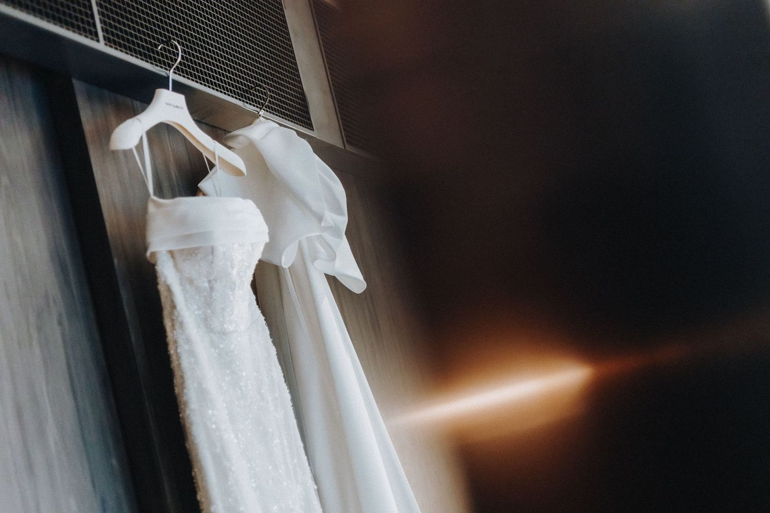 White wedding dress hangs elegantly on a hanger against a dark wall with dramatic lighting effect.