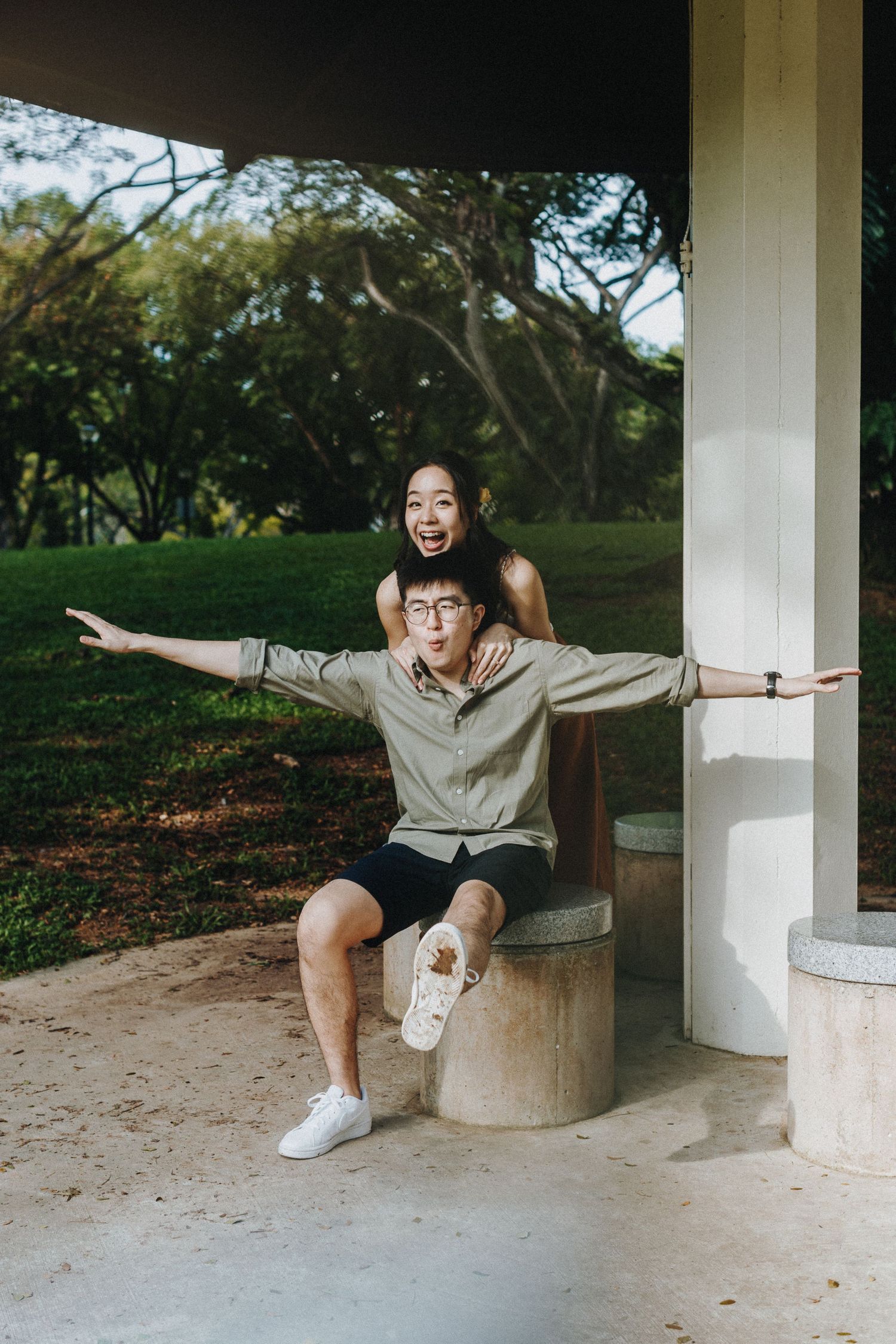 Playful couple posing underneath a white gazebo in a park with outstretched arms.
