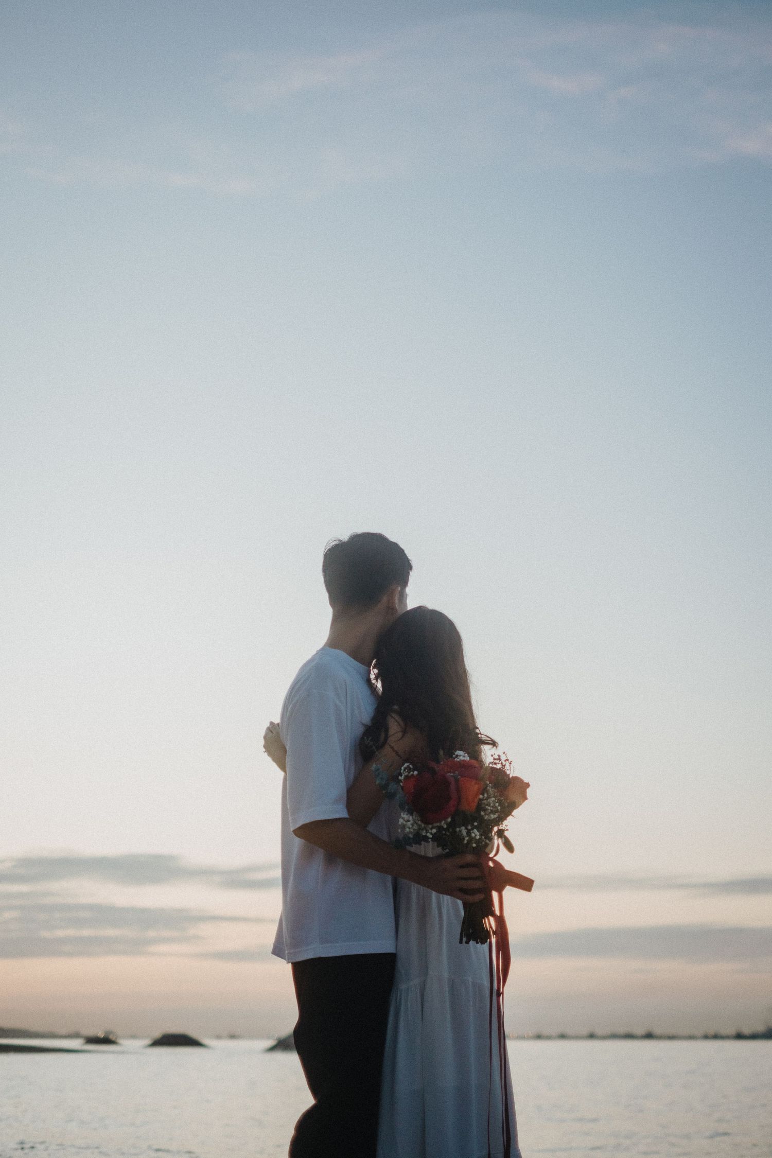 Couple embracing on beach during golden hour while holding bouquet of red roses.