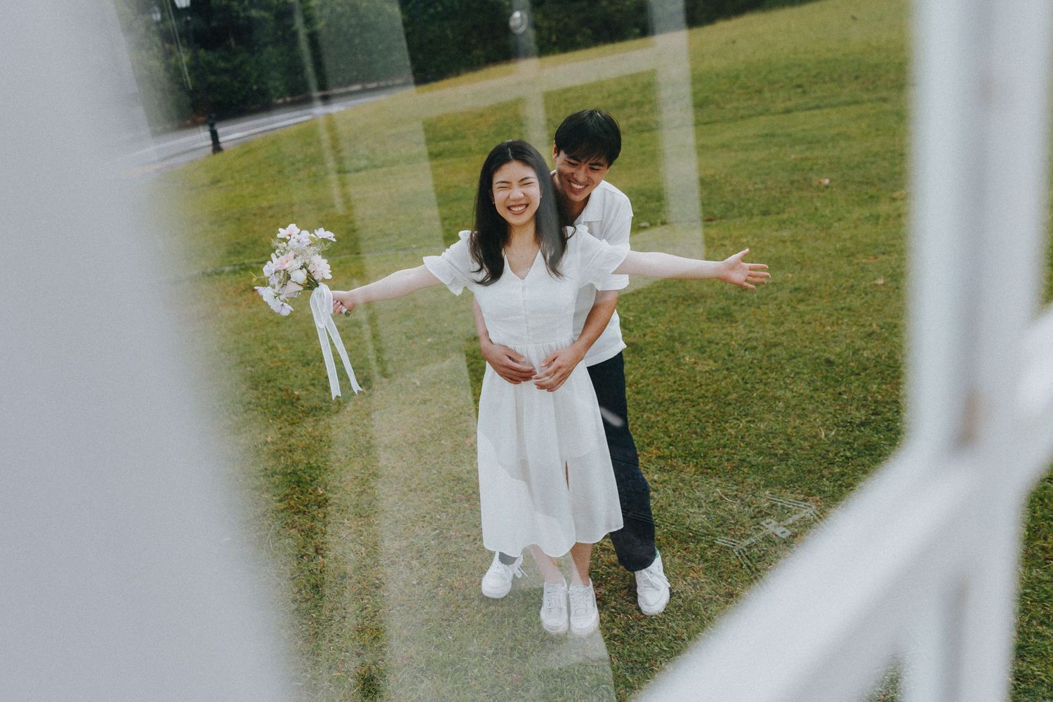 A joyful couple in white wedding attire embrace on a green lawn viewed through a white fence.