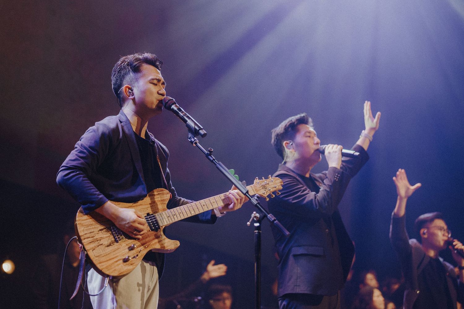 Musicians perform on stage with a guitarist playing and a singer gesturing under dramatic purple stage lighting.