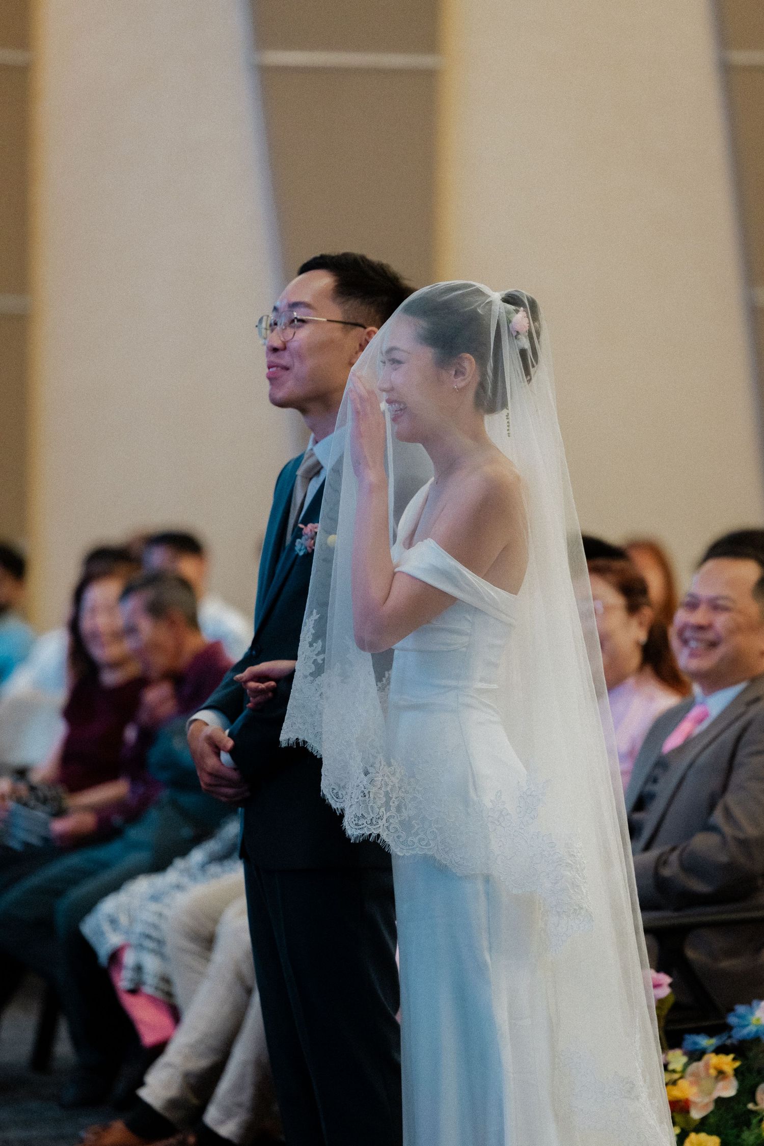 Emotional Asian bride wiping tears beside groom during wedding ceremony, seated guests smiling in background at elegant venue.