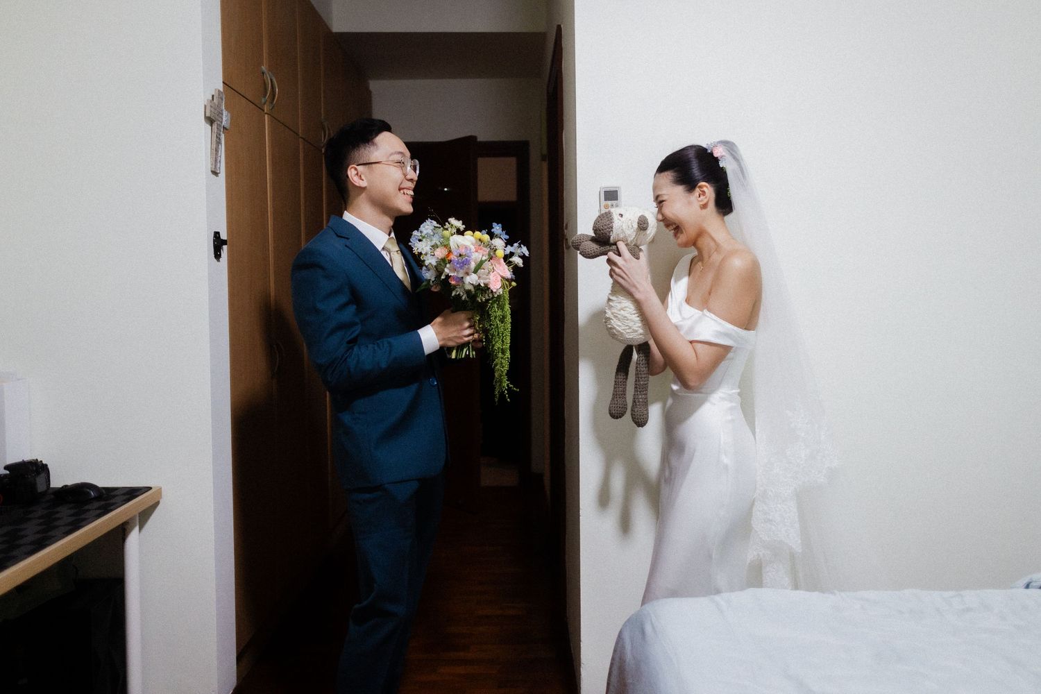 Bride in white off-shoulder wedding dress shows groom a stuffed toy as he holds a colorful bridal bouquet.