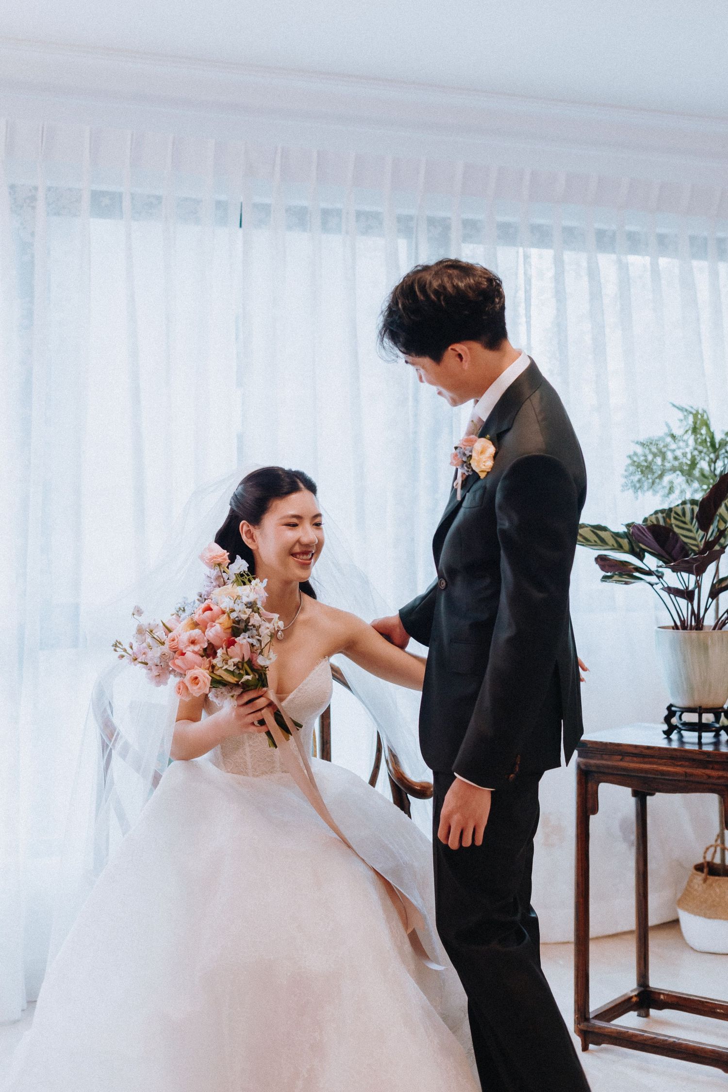 Bride in white strapless gown holding floral bouquet smiles at groom in black suit near sheer curtains.
