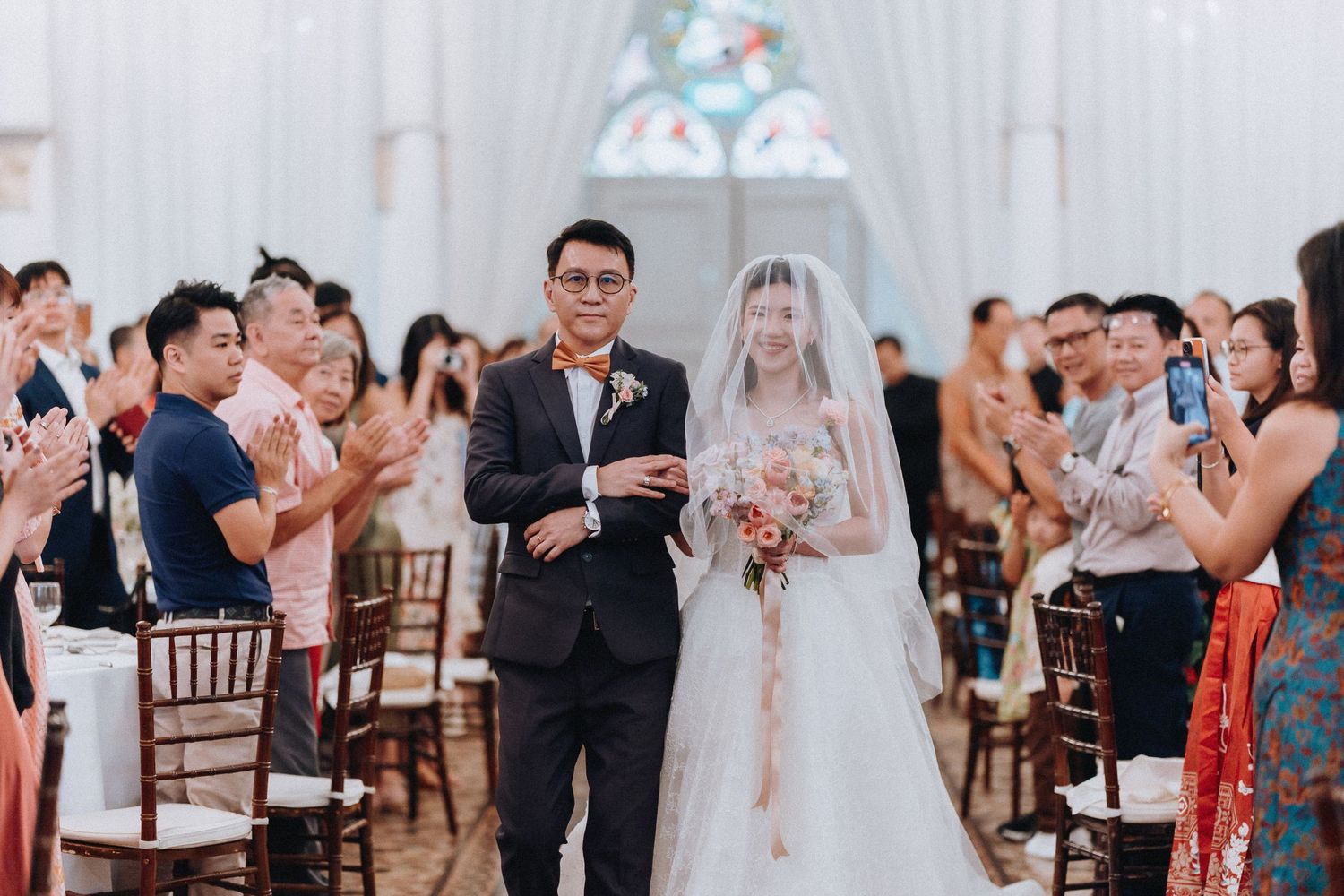 Bride in white gown walks down chapel aisle with escort as guests applaud during elegant wedding ceremony.