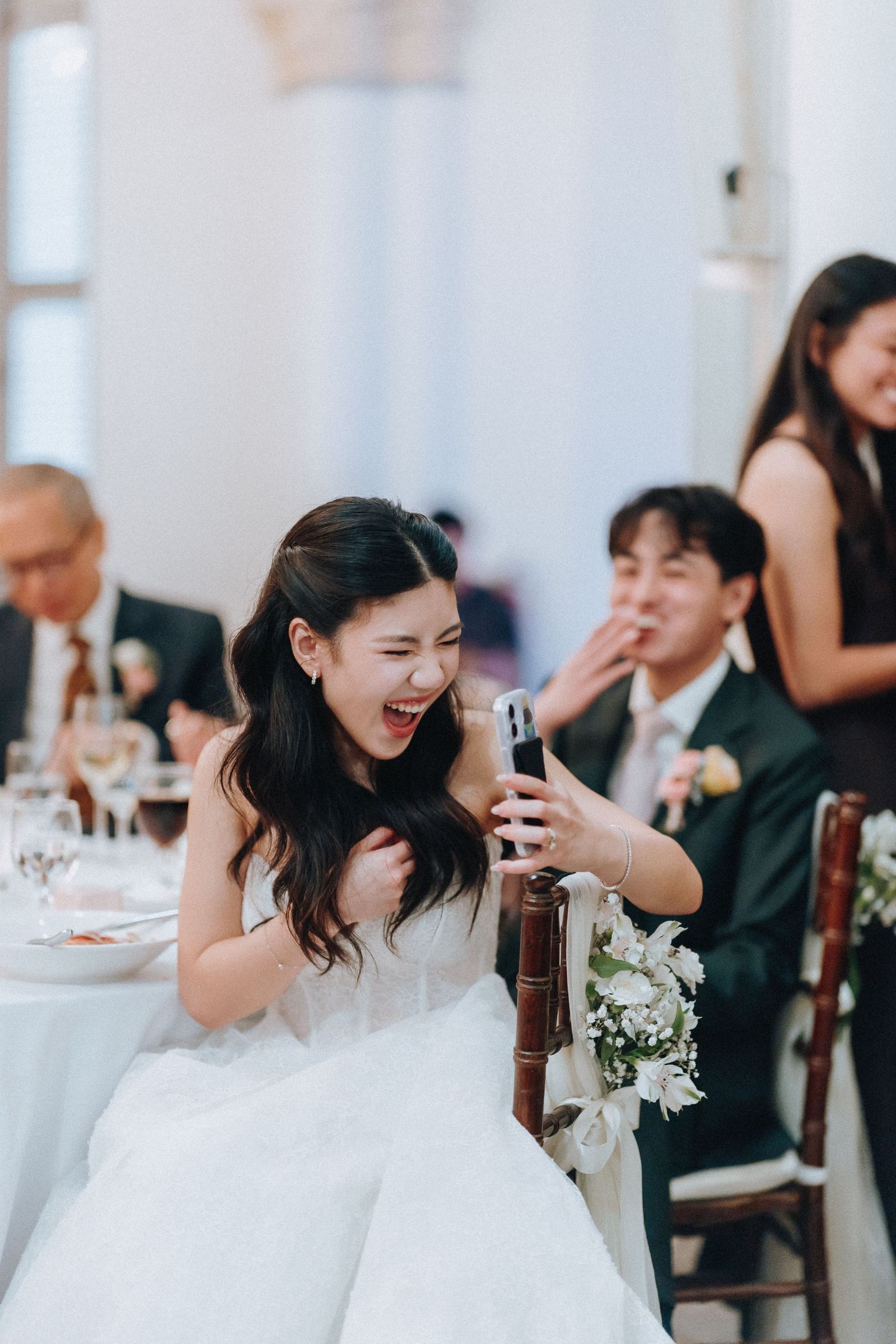 Joyful bride in white gown reacts with excitement while looking at her phone during wedding reception dinner.