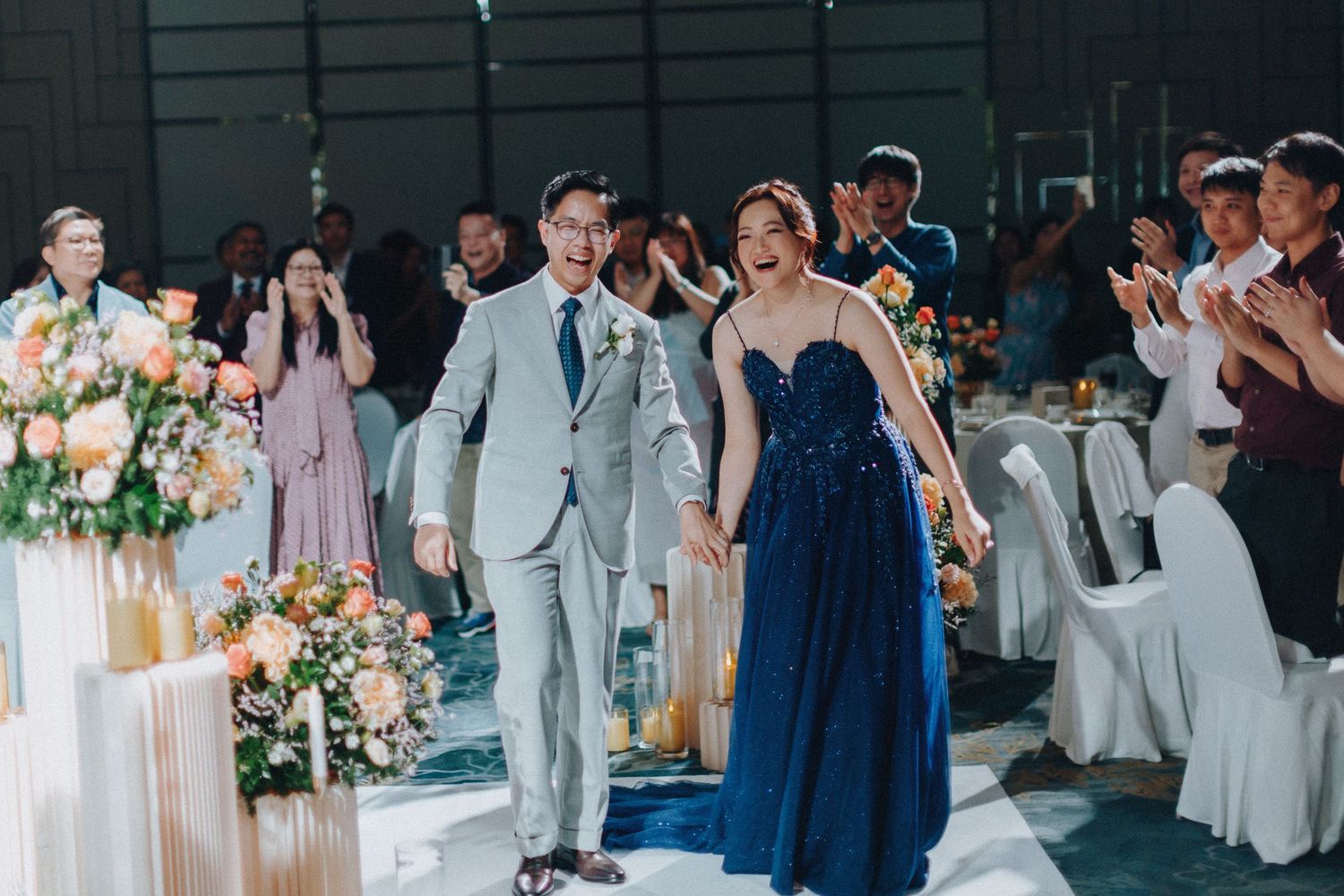 Wedding party in light gray suits and navy dresses dancing and celebrating at an indoor reception venue.