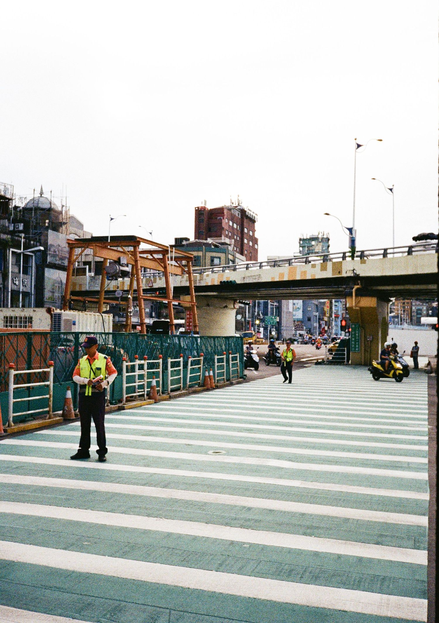 A striped pedestrian walkway with green and white stripes leads through a city area with buildings and railings visible.