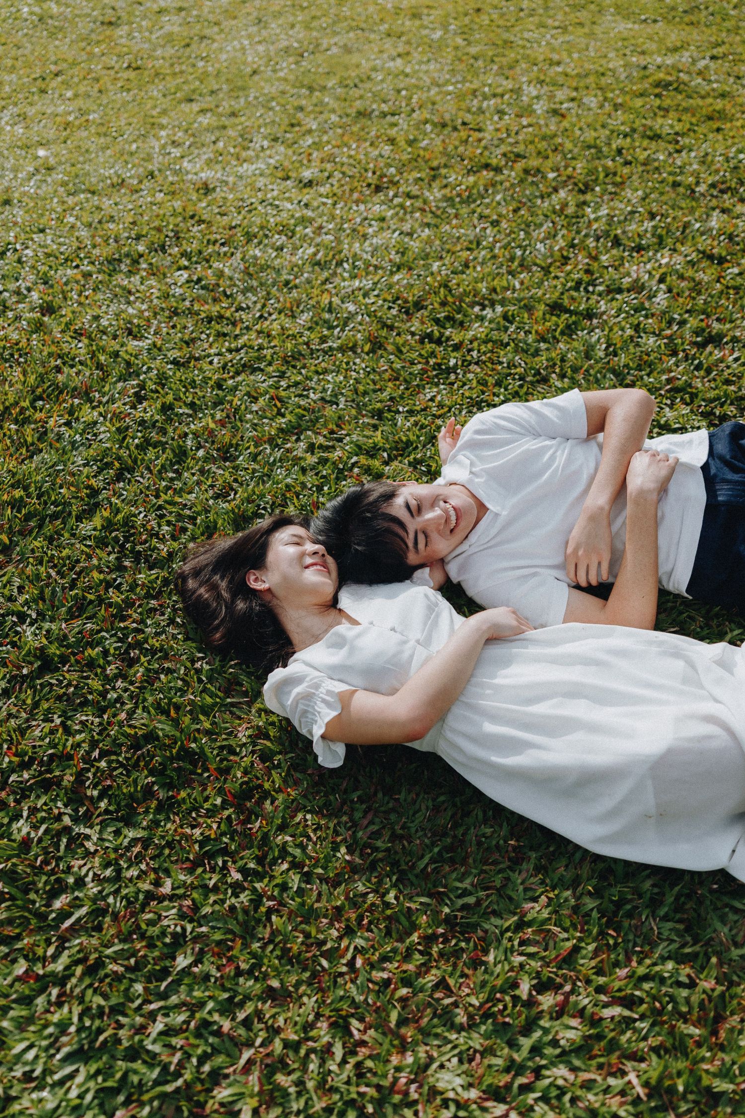 A couple in white outfits lying on grass and sharing a joyful moment together.