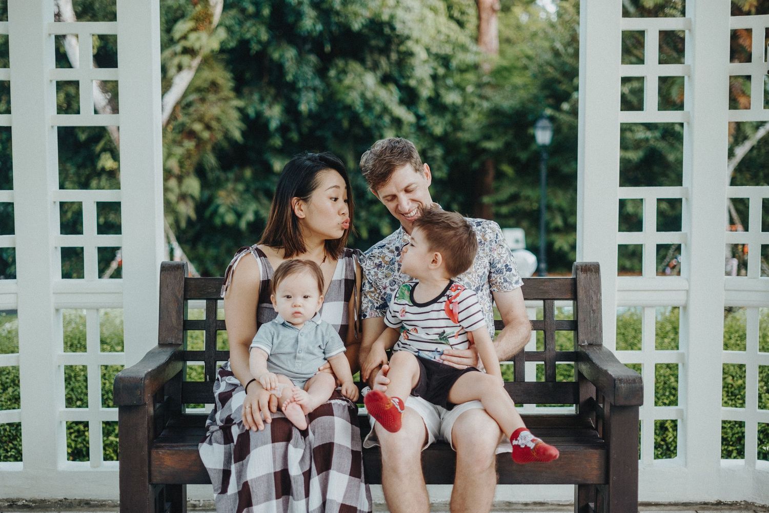 Family sits together on wooden bench in garden setting beneath white lattice arbor on sunny day.