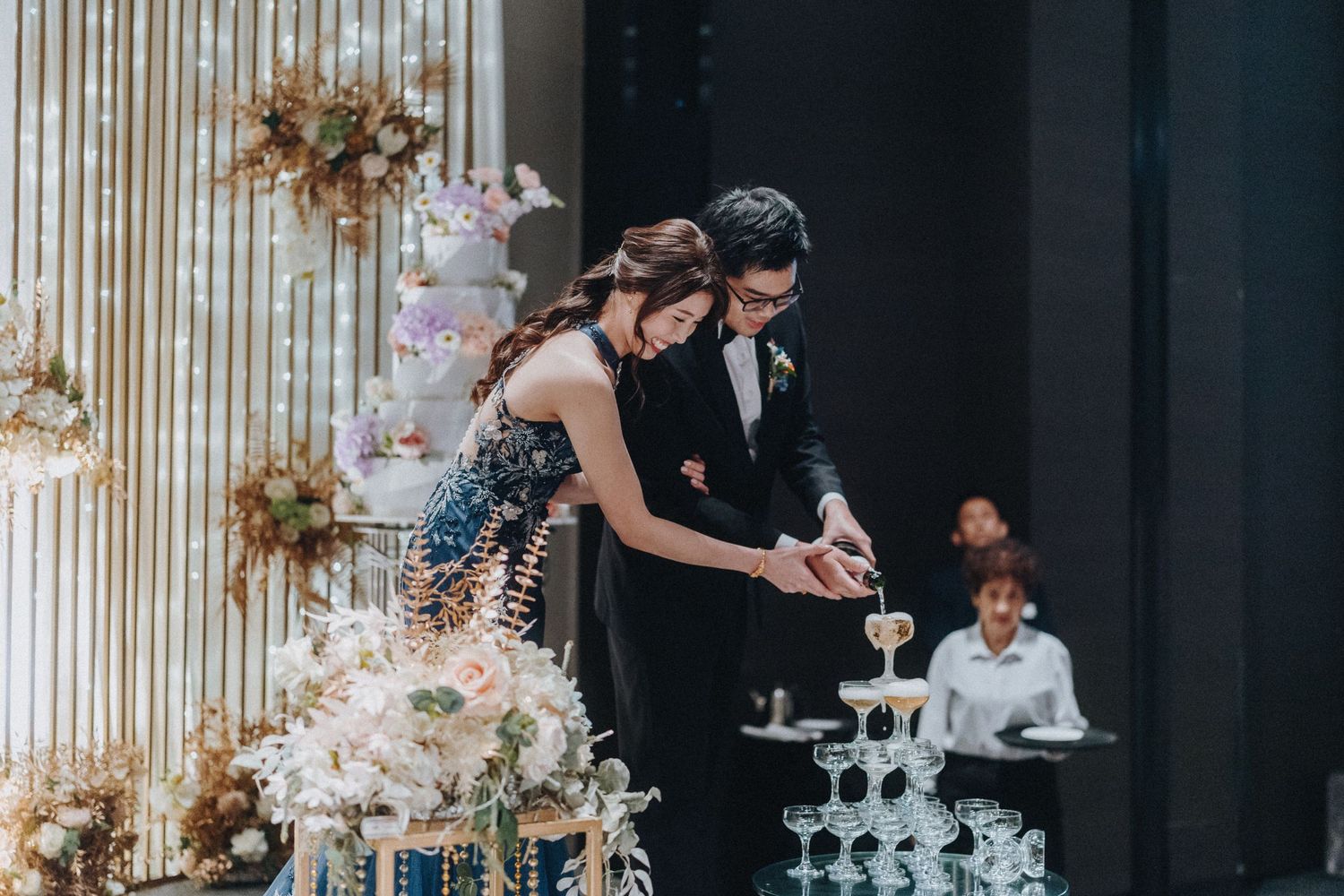 A couple in formal attire enjoys a private moment during a wedding reception with decorative lighting in the background.