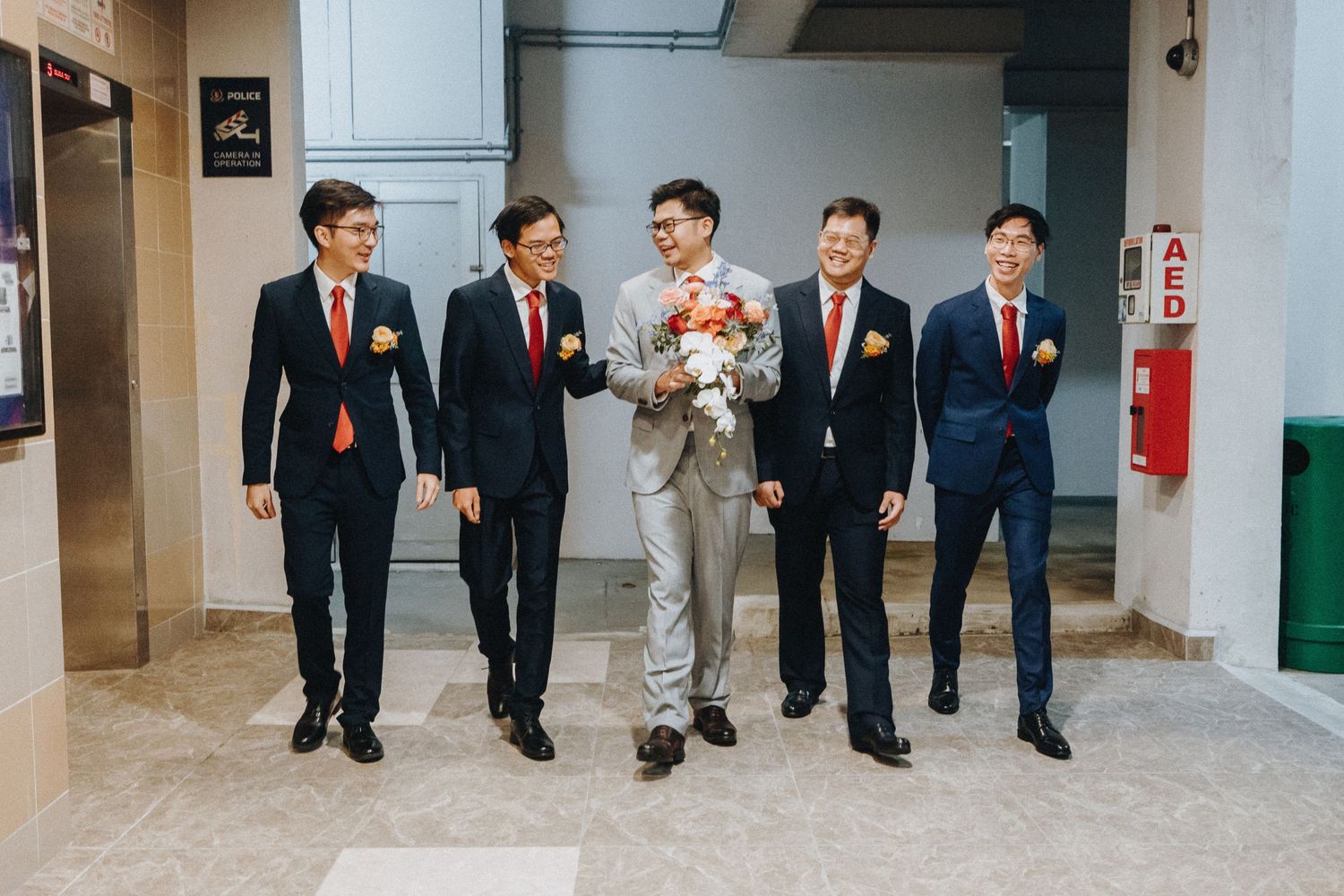Groomsmen in dark suits walk together indoors, with one in gray holding a pink and white floral bouquet.