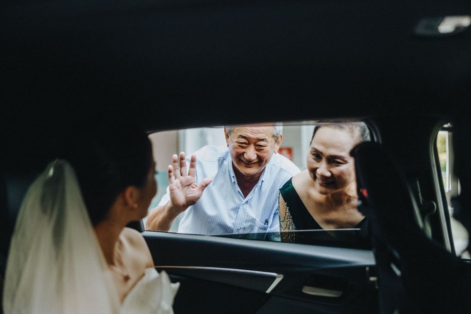 Two people wave through a car window at a bride on her wedding day.