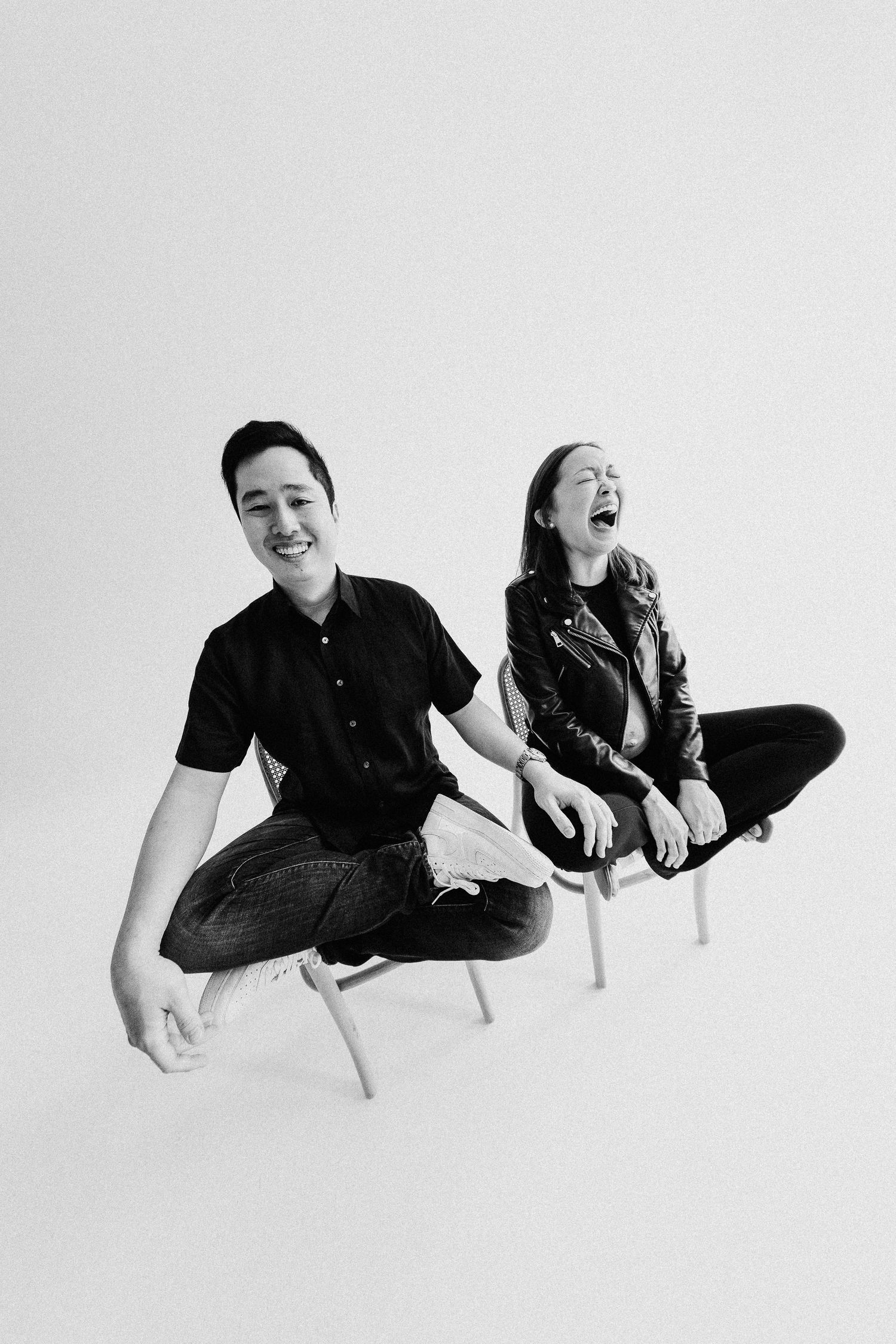 Two people sitting on chairs laughing together in a minimalist black and white studio setting.