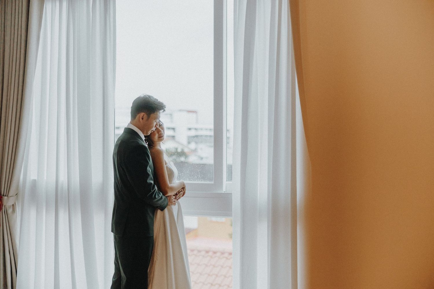 A romantic wedding moment showing a couple in formal attire standing together by a large window with sheer curtains.