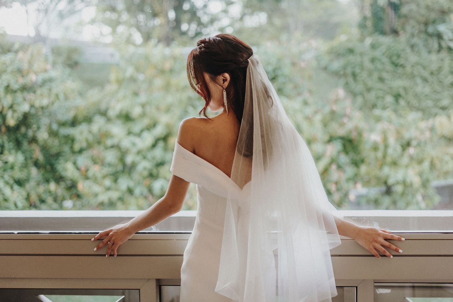 A bride in an elegant off-shoulder white gown and long veil stands by a window with her back to the camera.