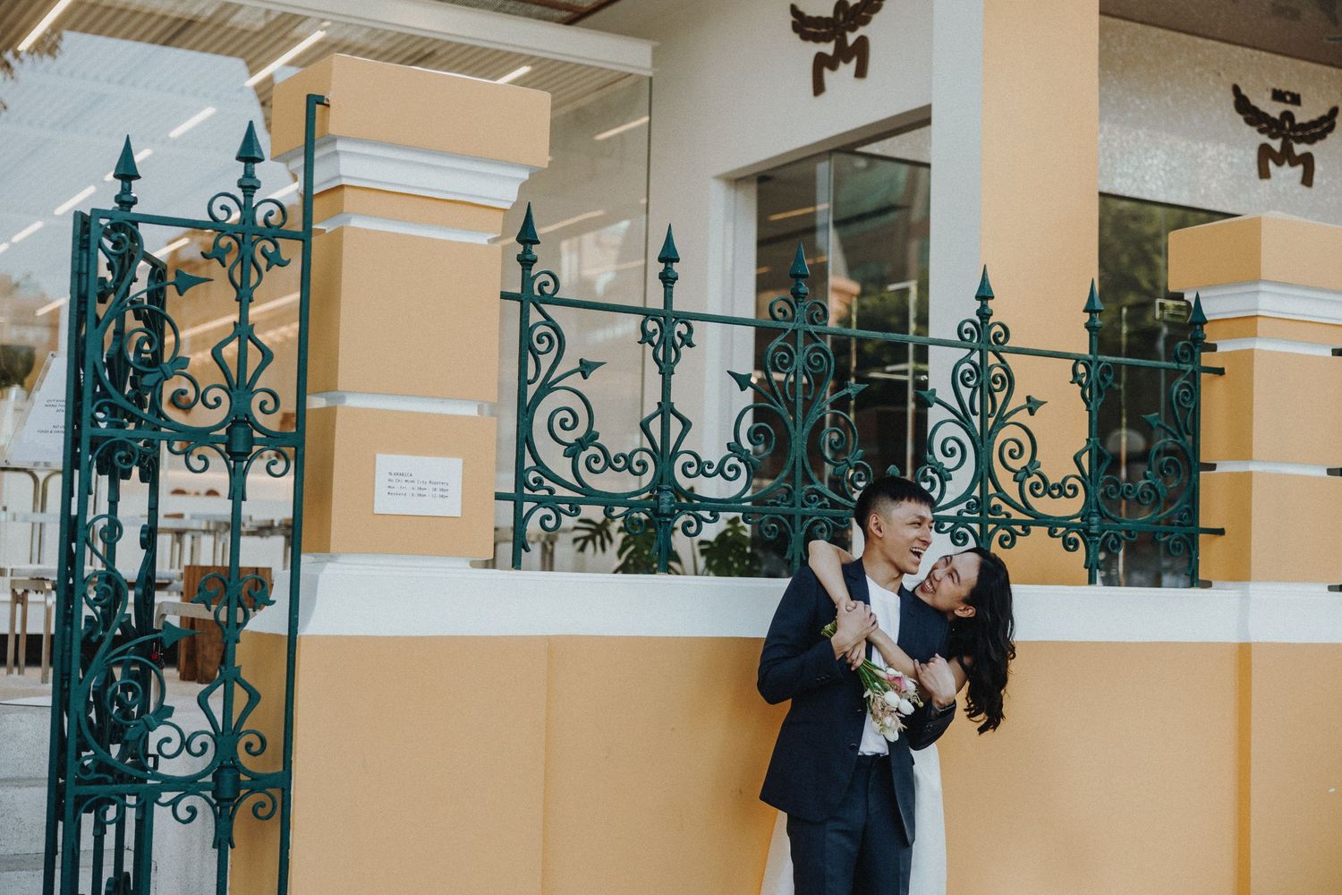 A couple embraces next to an ornate turquoise wrought iron gate against a yellow colonial-style building.