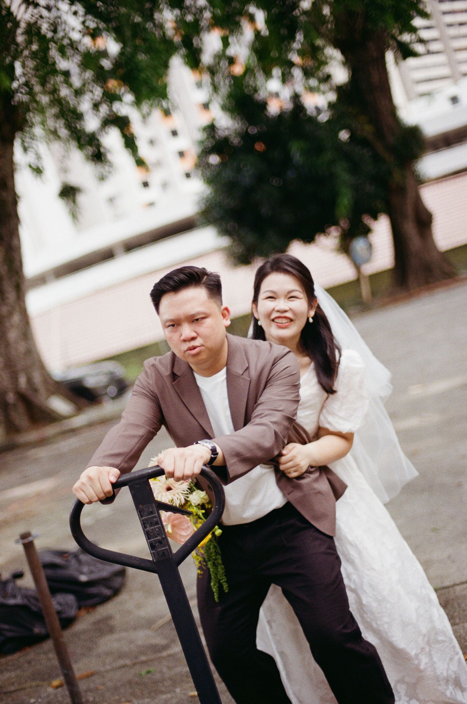 A couple in wedding attire share a lighthearted moment while sitting outdoors under trees.