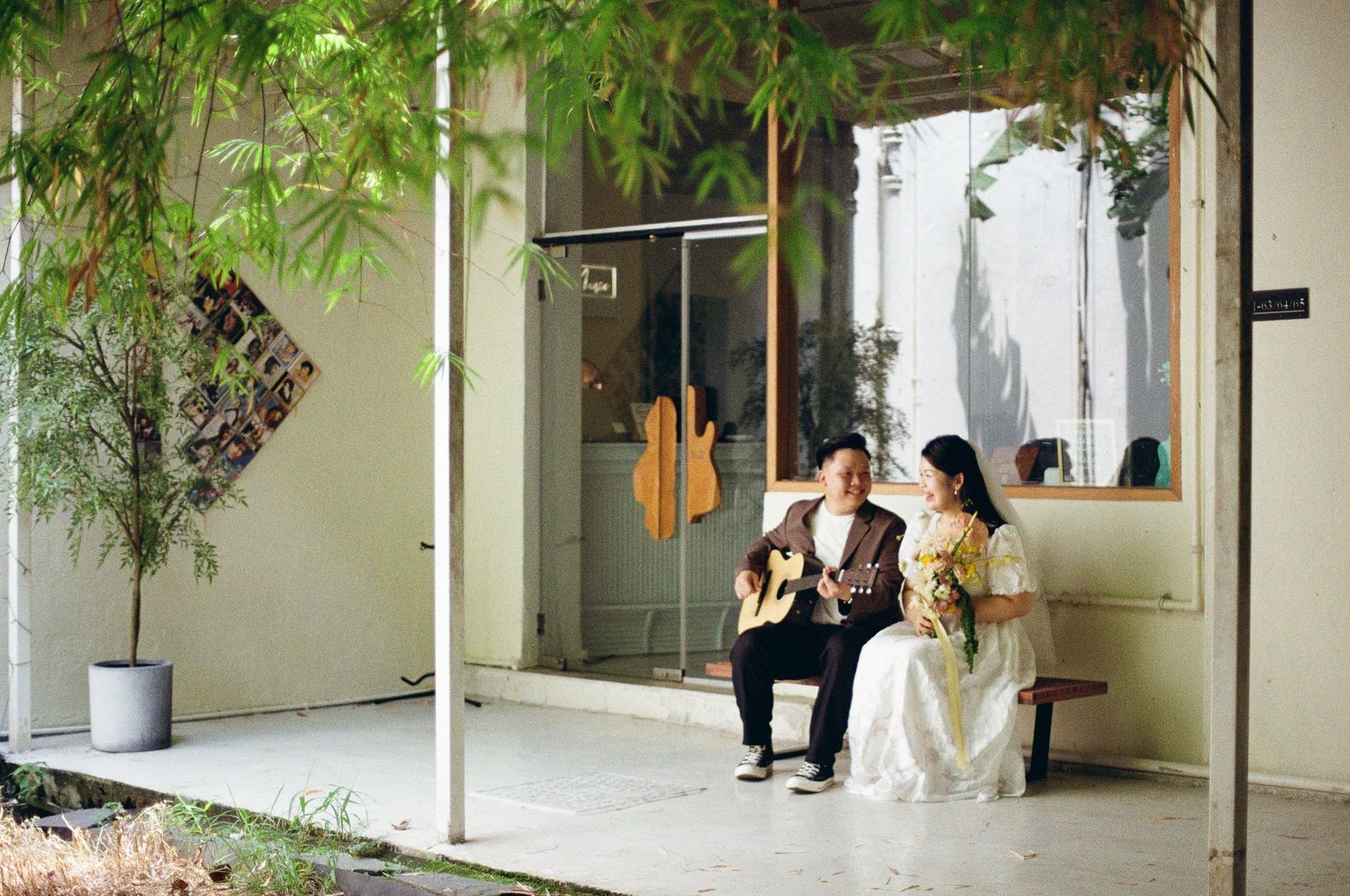 A couple sits together on an outdoor bench under a covered patio with green plants and white walls.
